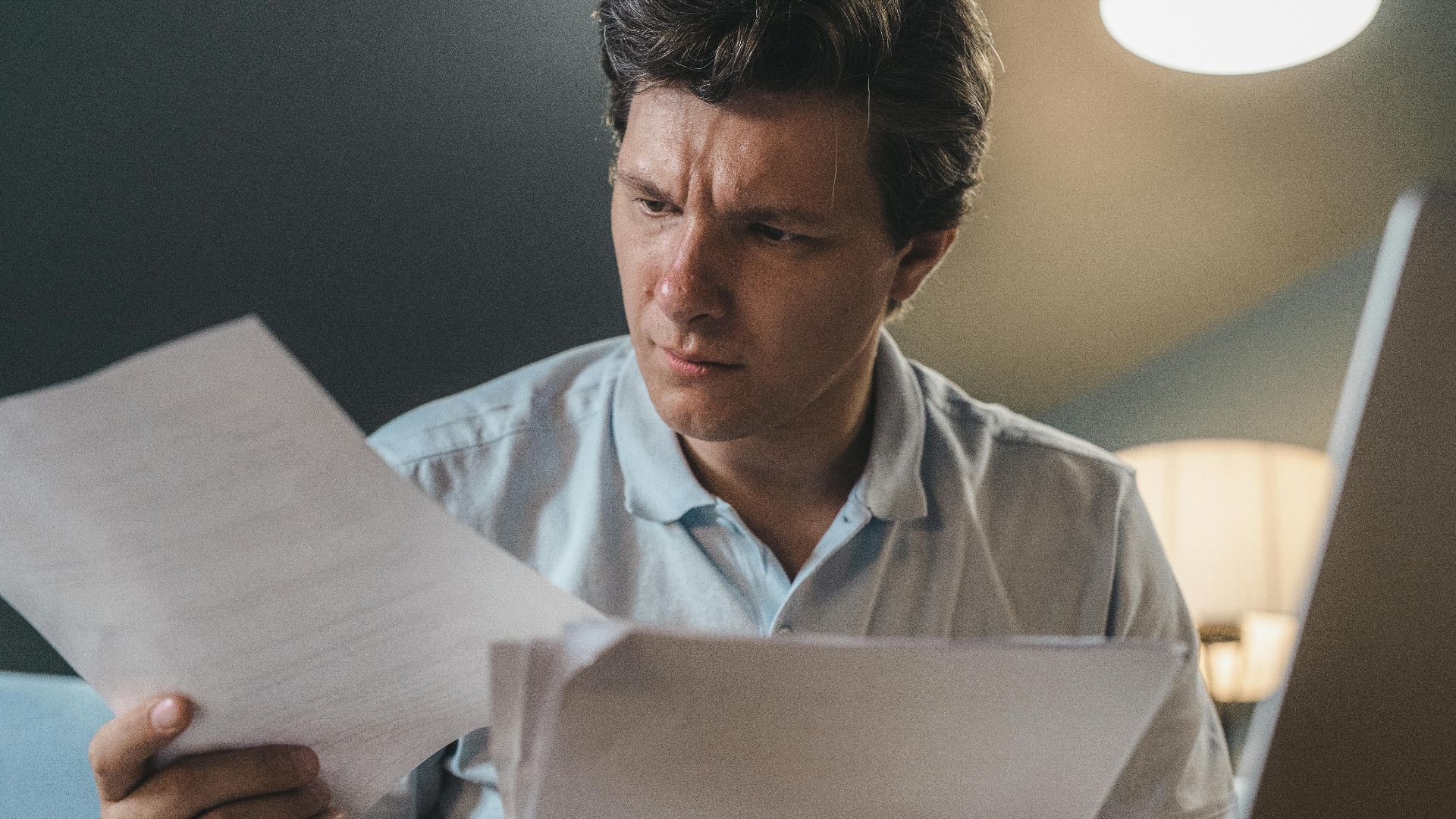 A man intently examines papers, seated indoors under warm lighting, focusing on his work.