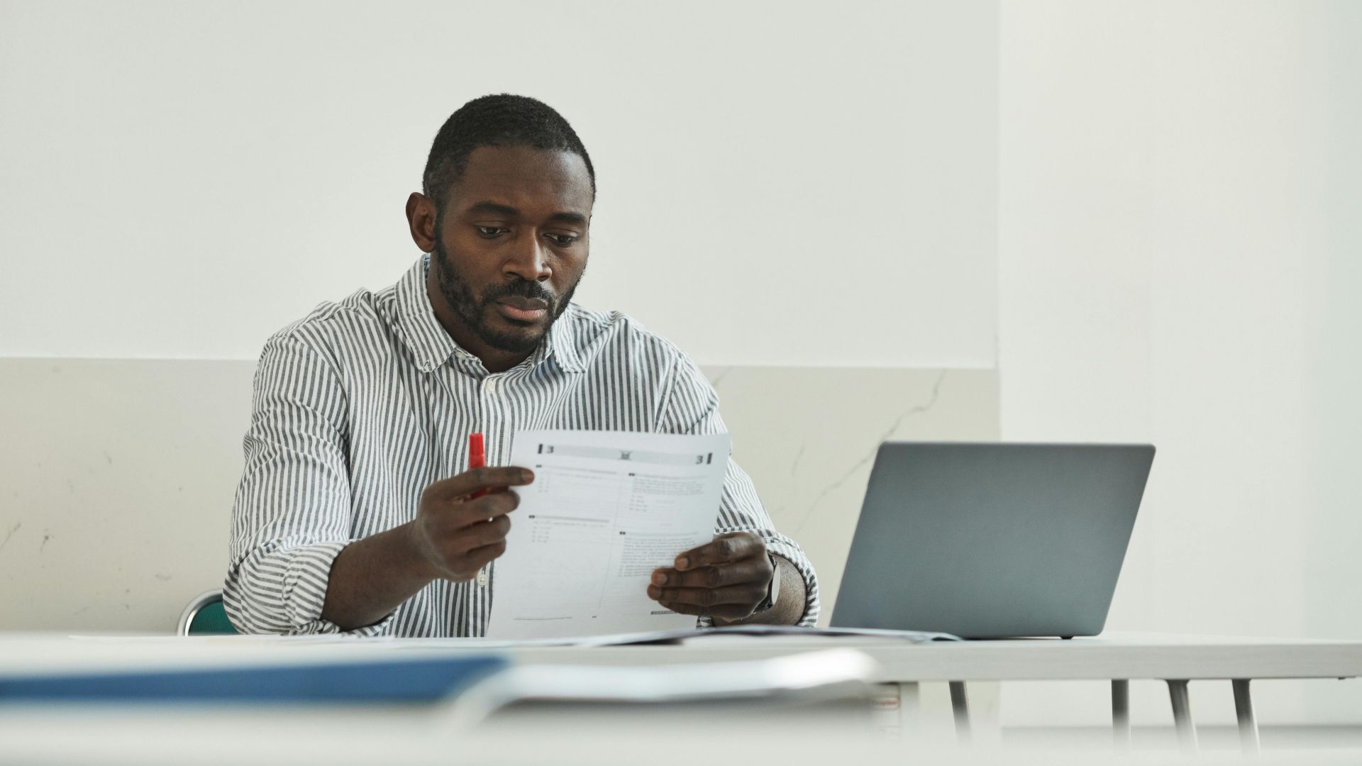 An adult man attentively grading test papers at his workspace with a laptop.