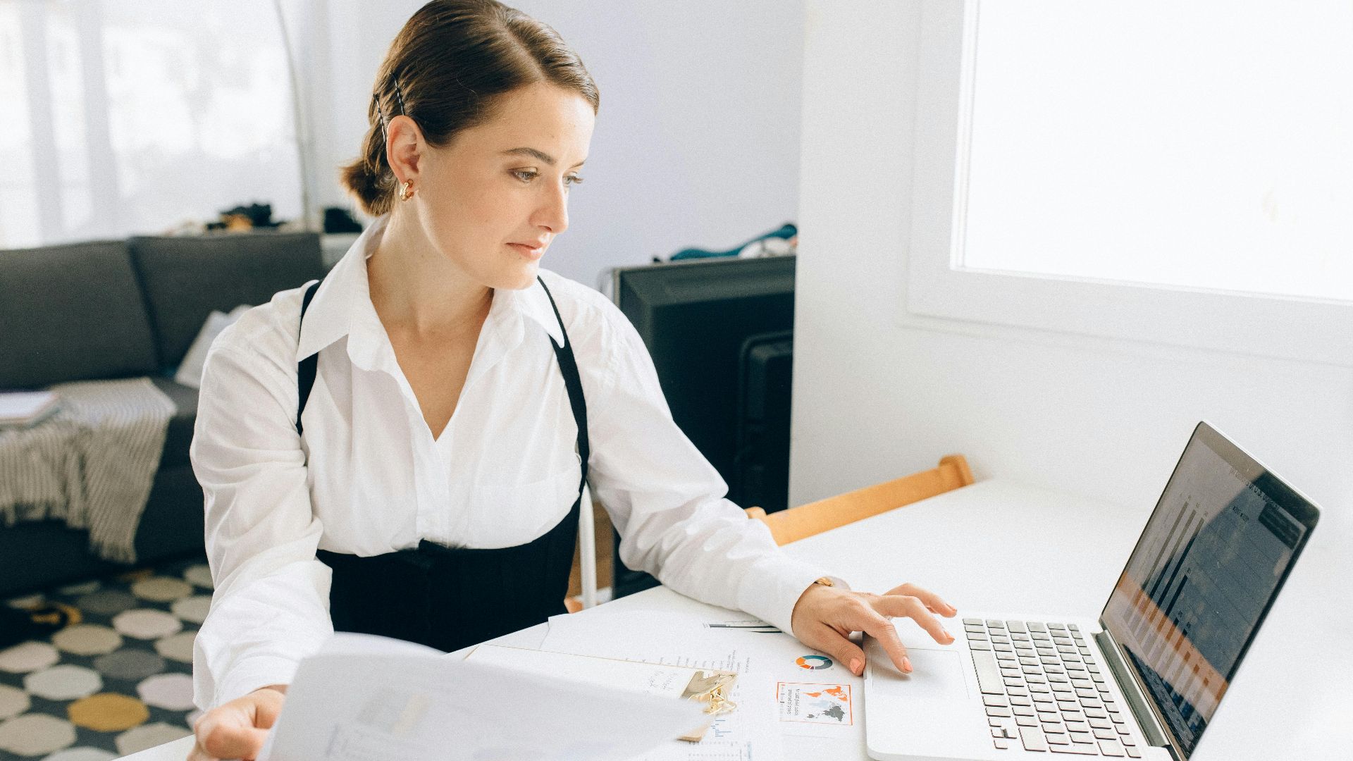 Woman at desk reviewing business reports on laptop, holding papers in a modern office setting.