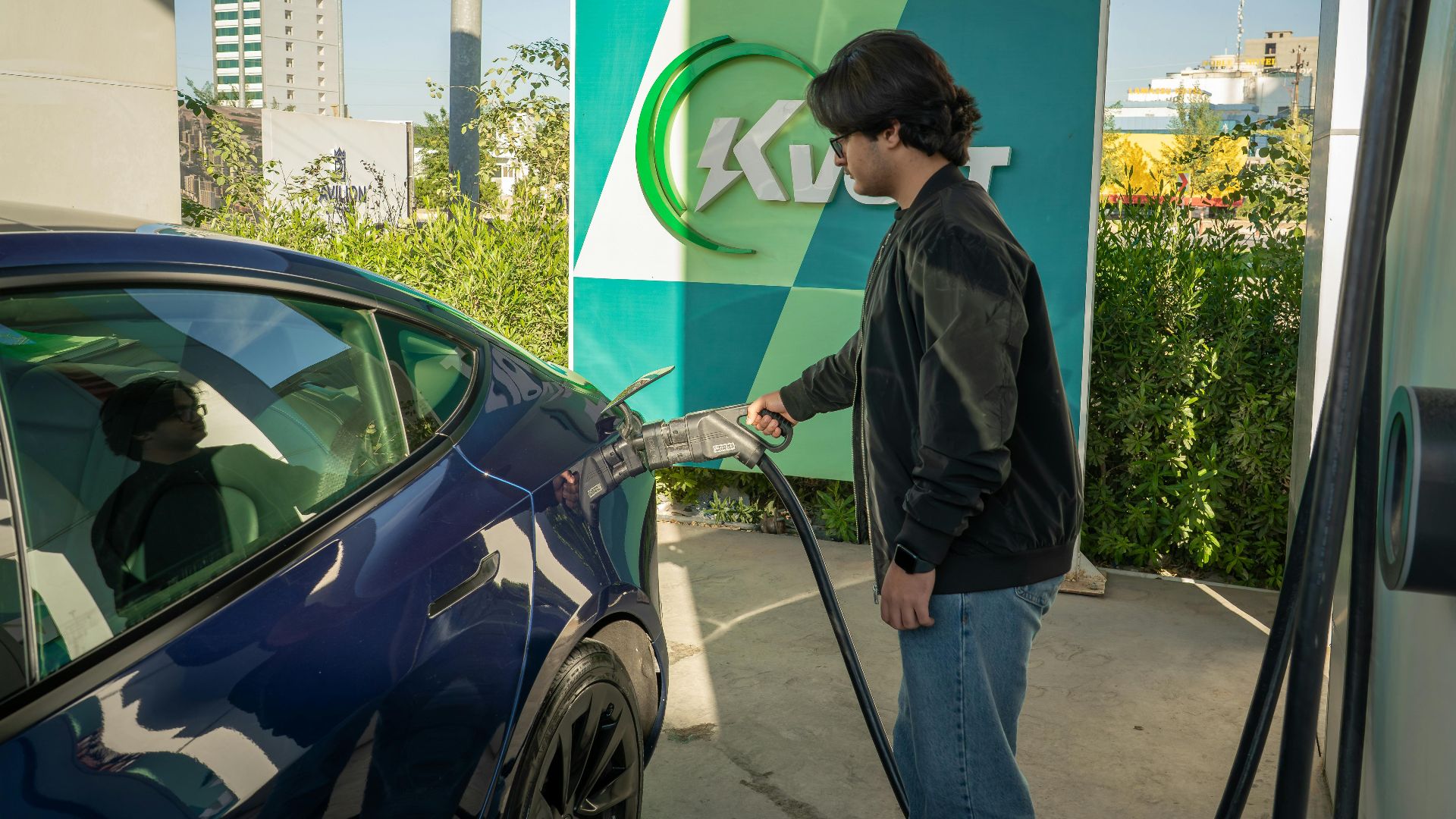 Young man charging a Tesla Model 3 at an outdoor electric vehicle station during the day.