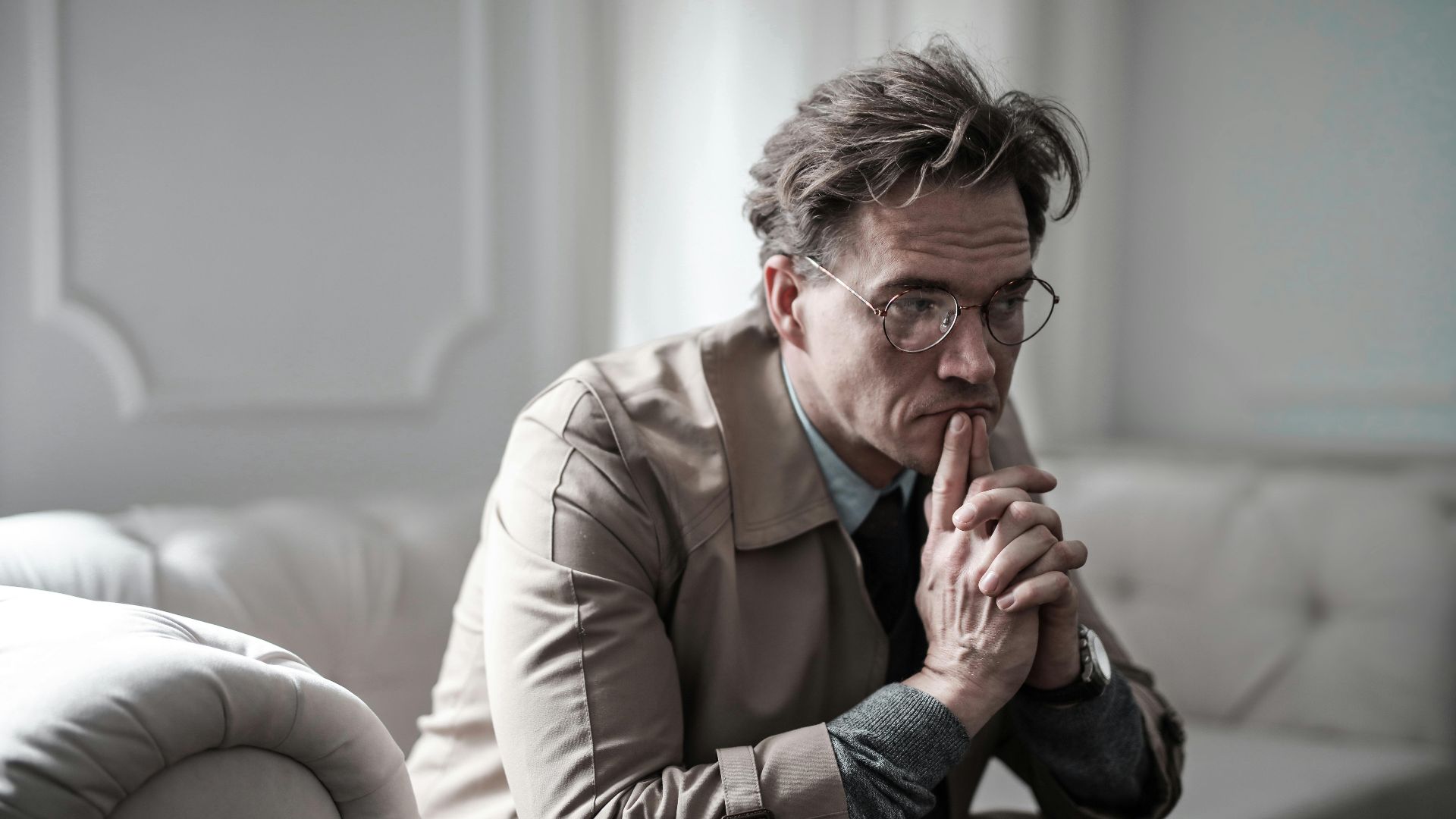 A thoughtful man wearing glasses sits on a sofa in a classic interior, lost in deep contemplation.