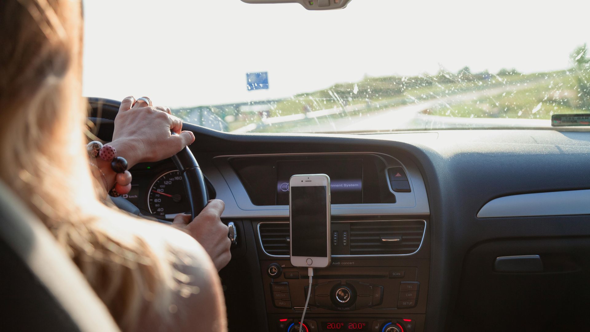 View from behind a woman driving a car on a sunny day with a phone mounted on the dashboard.