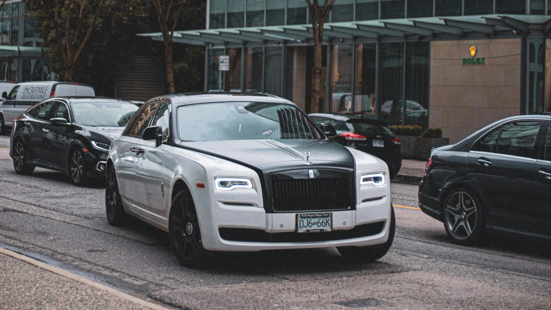 White Rolls Royce cruising through a city street with modern architecture and autumn foliage.