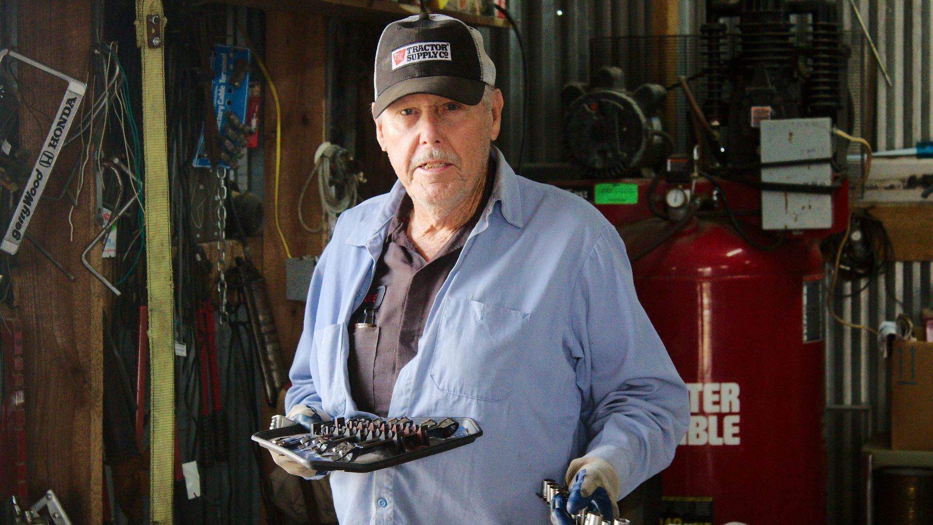Portrait of an older mechanic holding tools inside a workshop setting, surrounded by equipment.