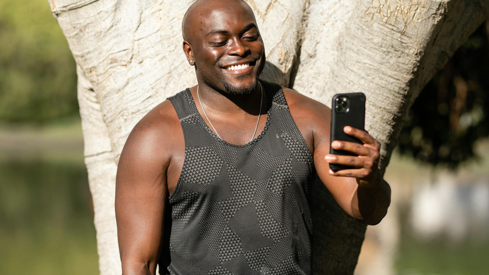 Man smiling while using smartphone near a tree in a sunny park.
