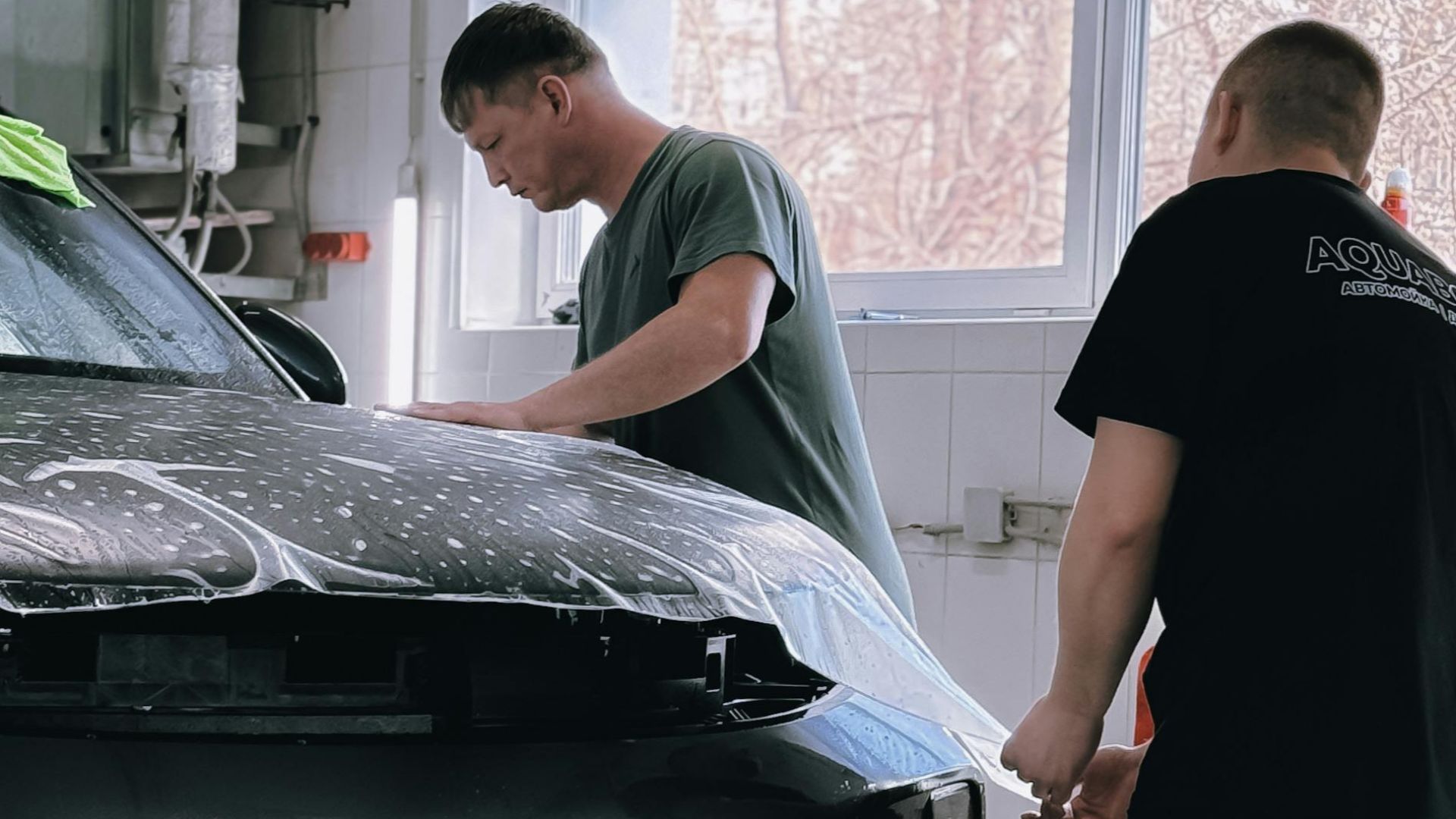 Two professionals applying protective film to a black car in an indoor workshop.