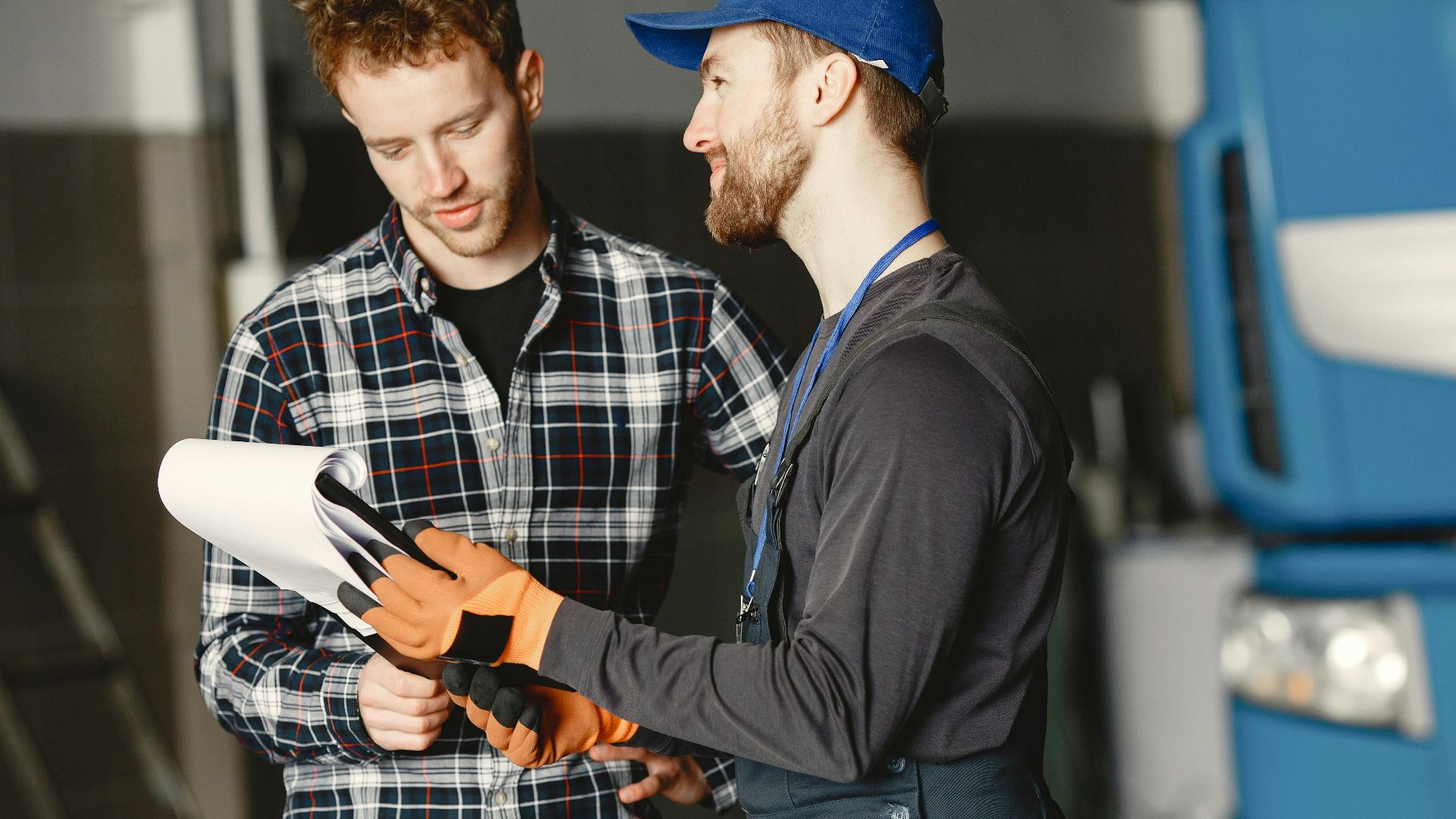 Mechanic in uniform consults with customer in garage setting beside a blue truck.