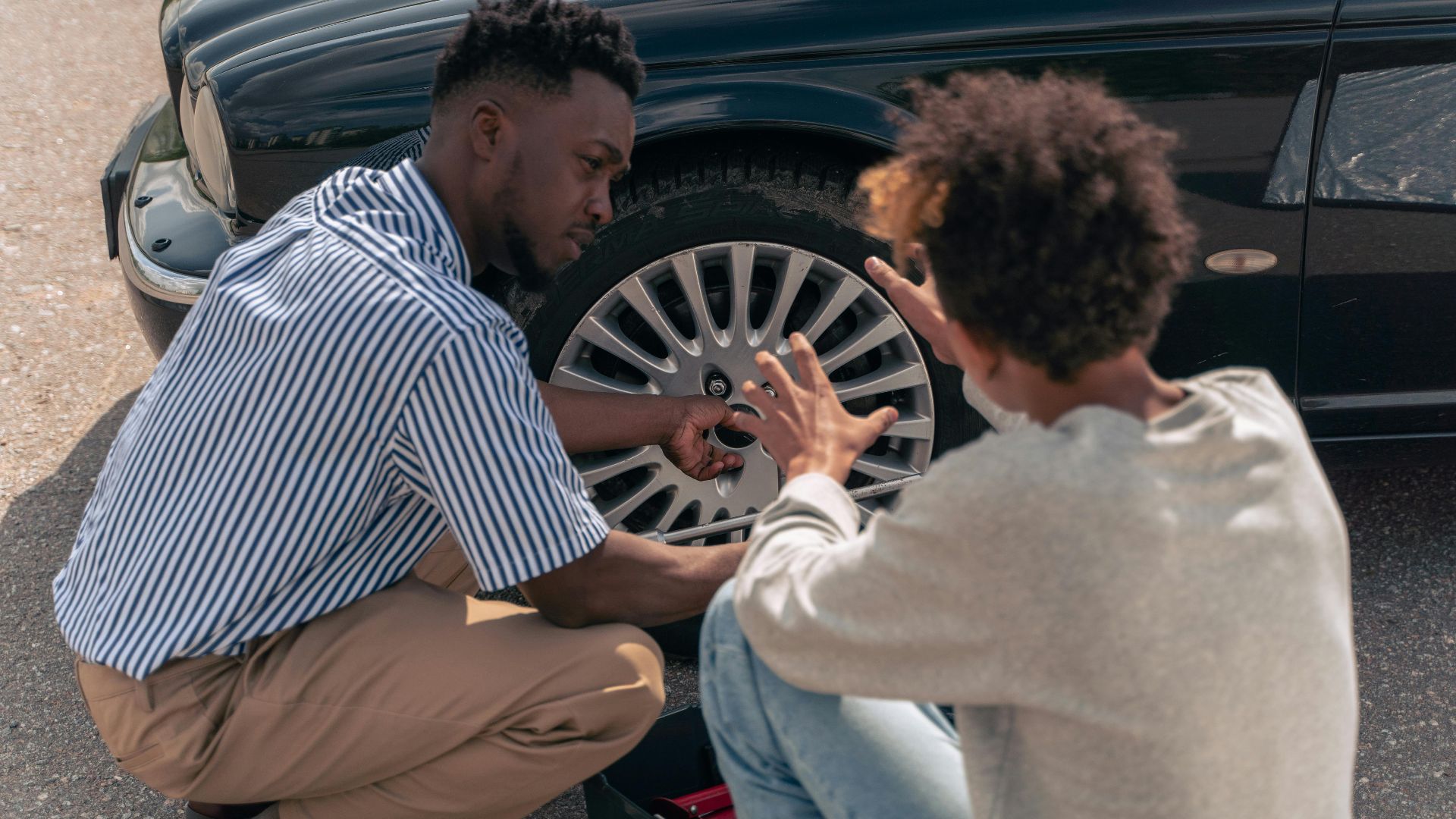 A father instructs his teenage son on how to change a car tire outdoors.
