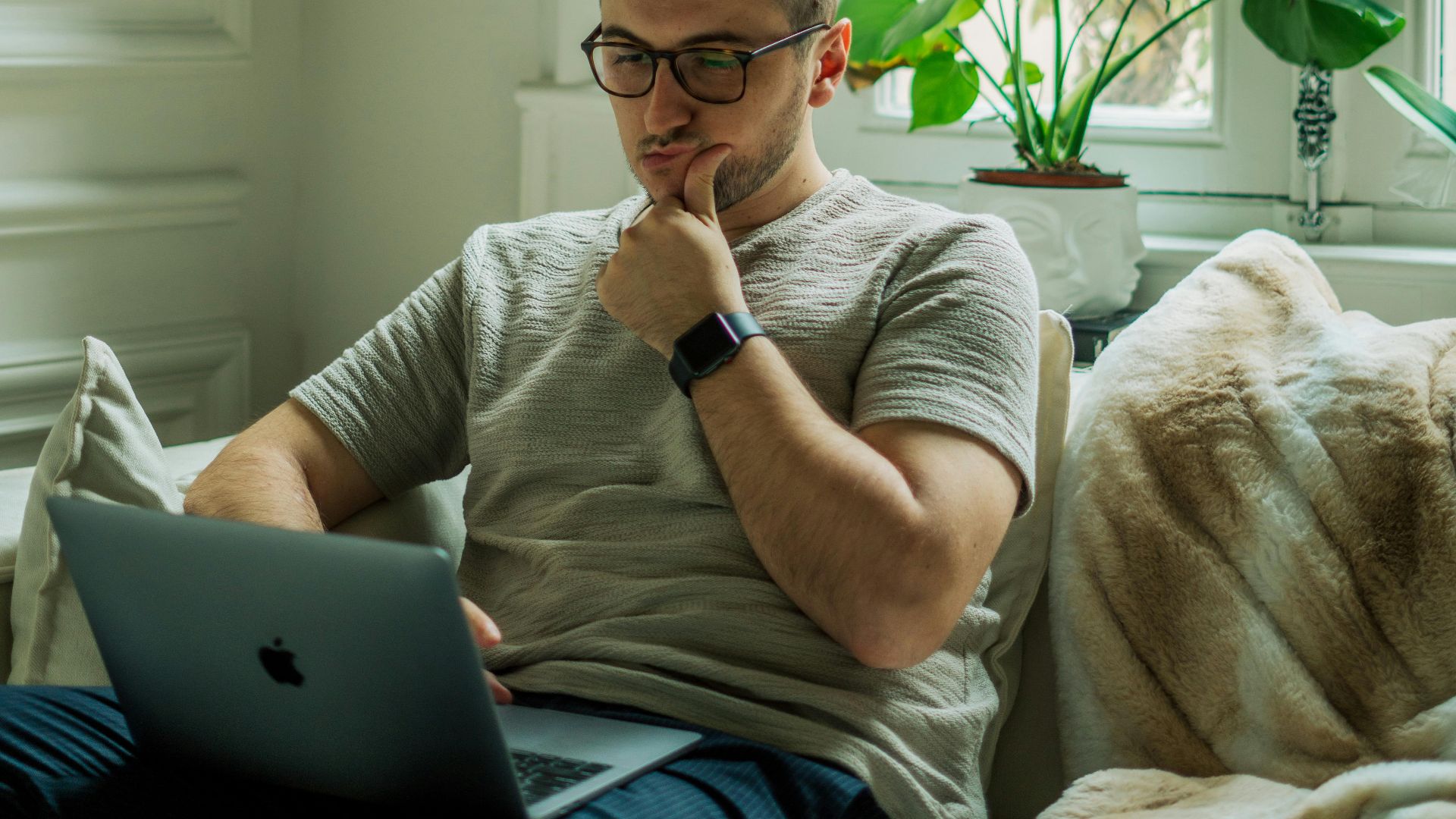 A man wearing eyeglasses using a laptop in a cozy home setting with natural light.