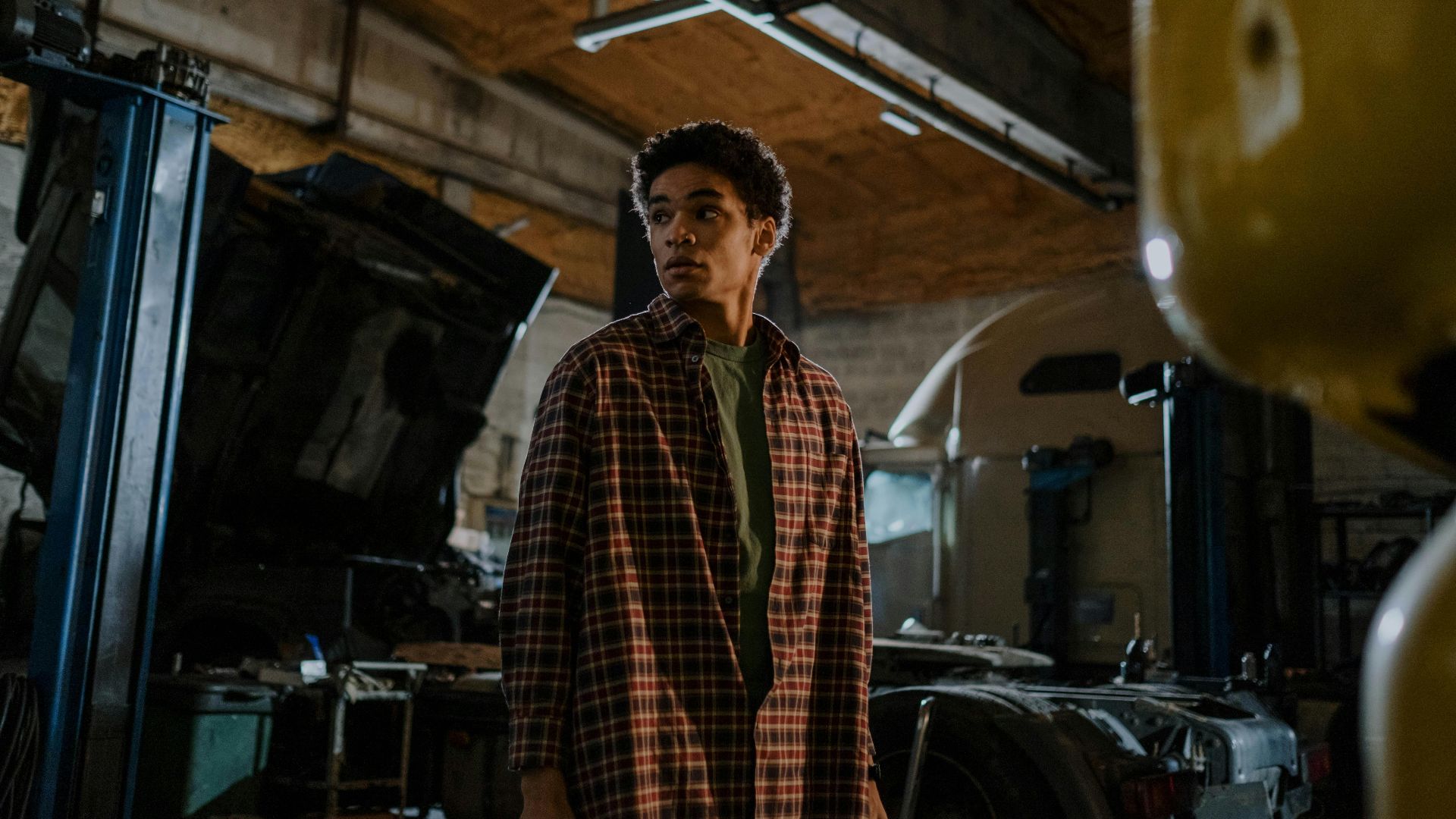 A young mechanic in a plaid shirt stands in a dimly lit auto workshop, deep in thought.