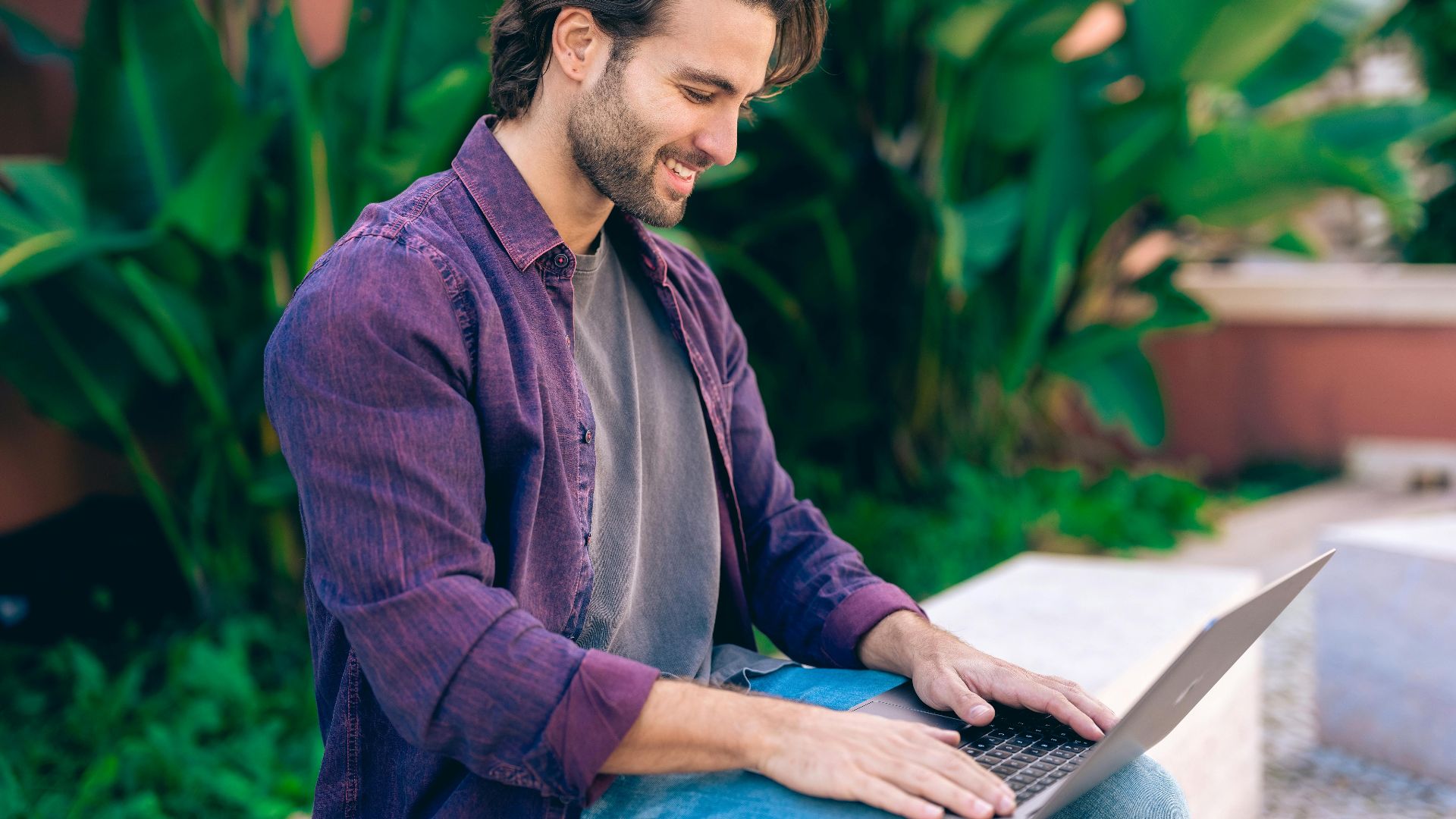 A smiling man in Lisbon using a laptop outdoors, surrounded by lush greenery. Perfect for lifestyle and remote work themes.