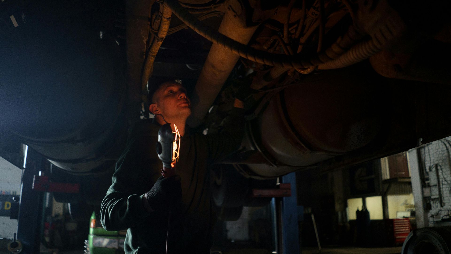 Mechanic examines vehicle underbody in dimly lit workshop, holding inspection light.