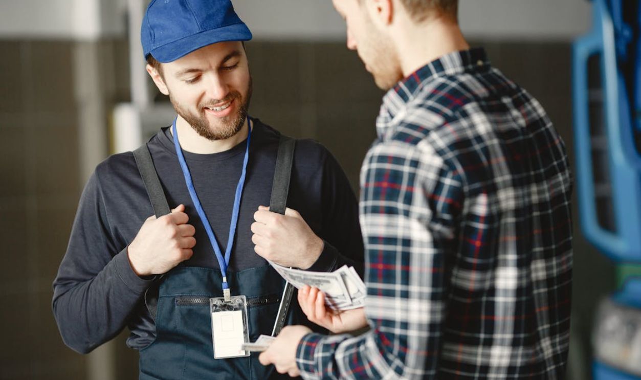 Man in Plaid Shirt Giving Money to Bearded Man in Blue Cap and Overall