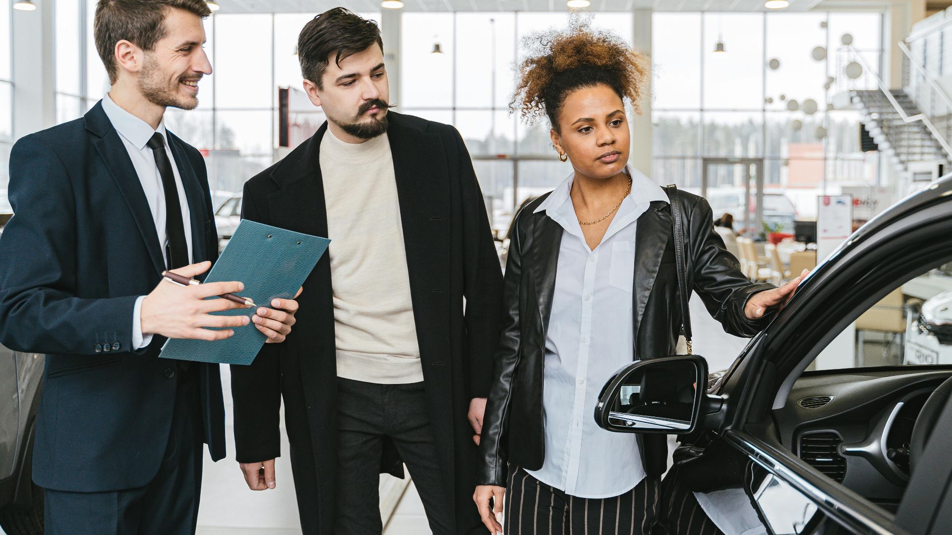 An interracial couple consults with a salesman at a car dealership, exploring vehicle options.