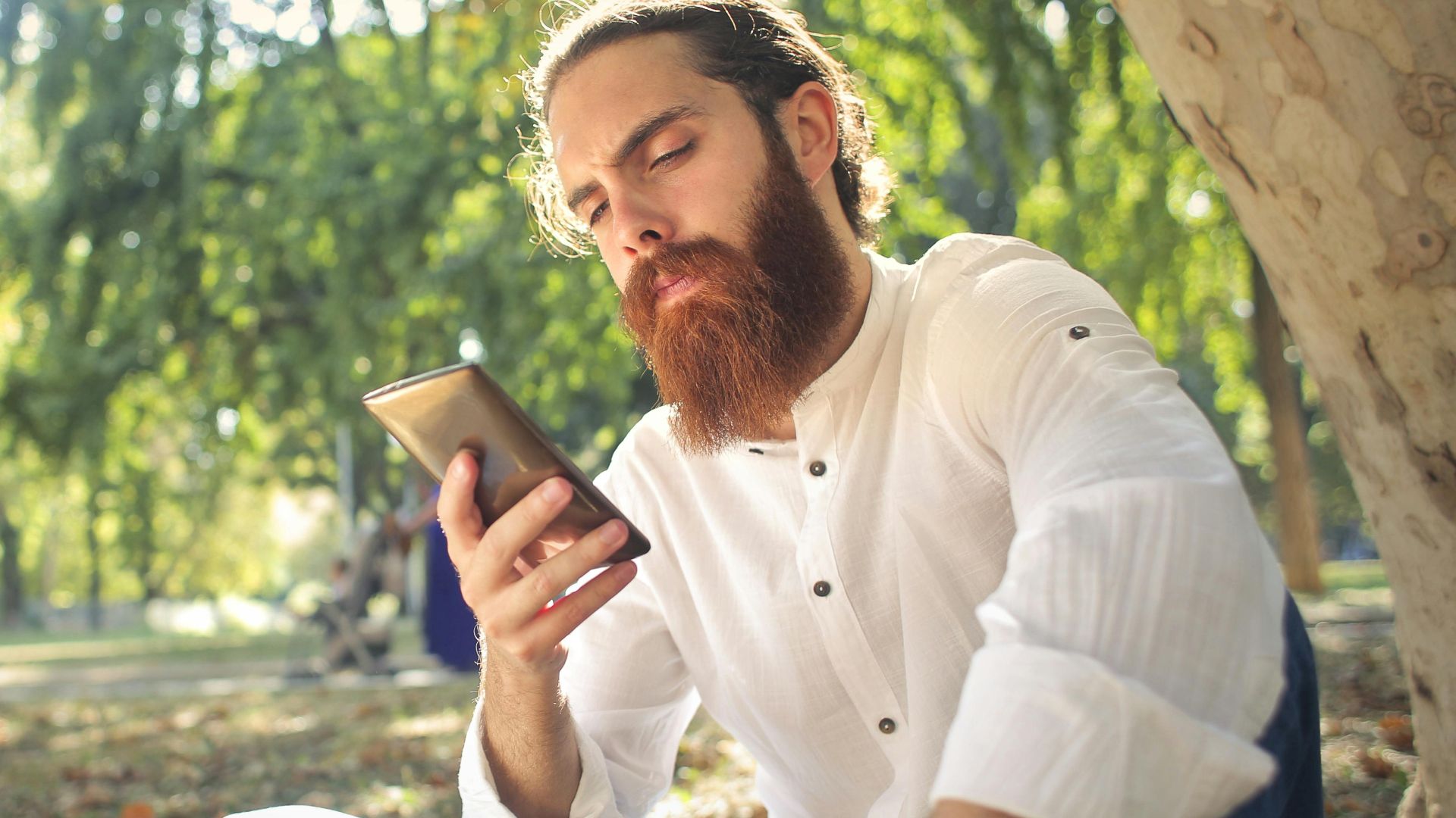 A bearded man in a white shirt uses a smartphone while sitting outdoors in a park.