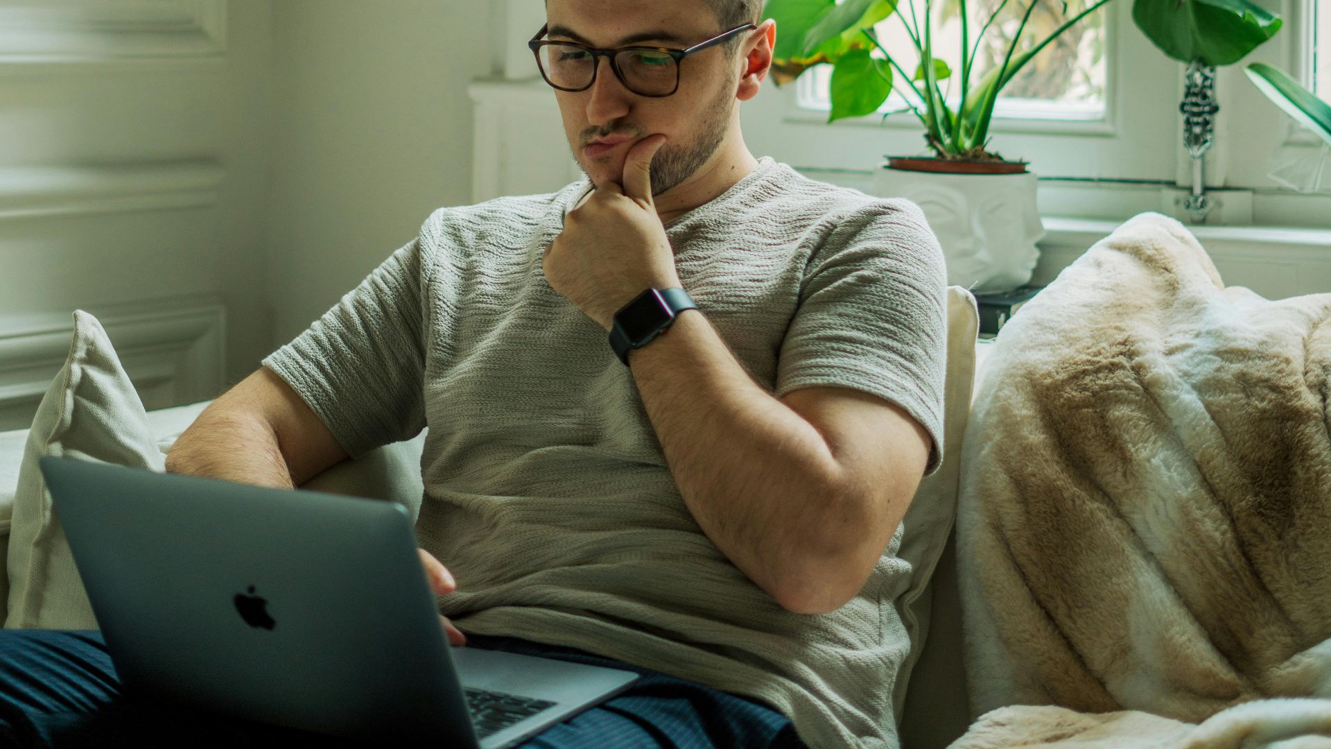 A man wearing eyeglasses using a laptop in a cozy home setting with natural light.