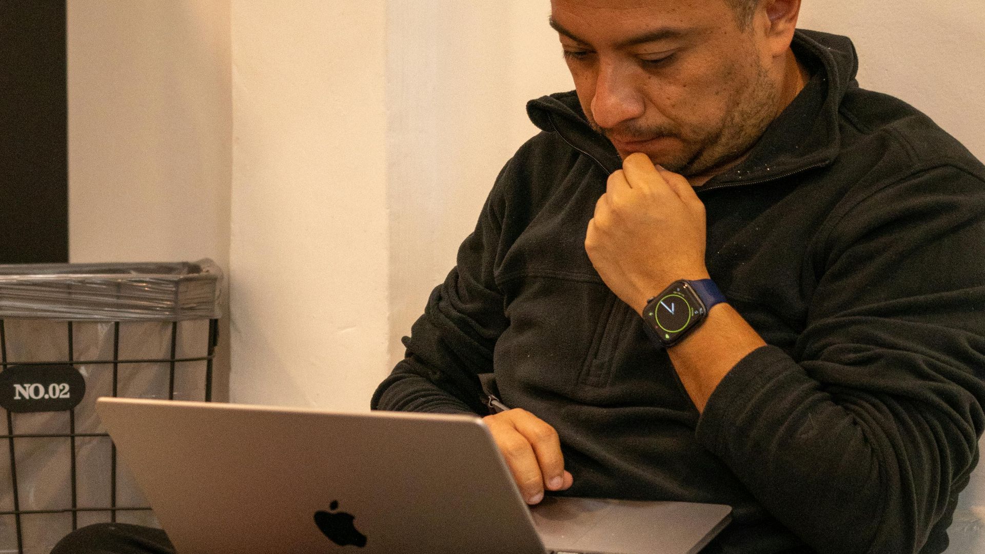 Man sitting cross-legged indoors, focused on his laptop. Casual setting, contemplating or working.