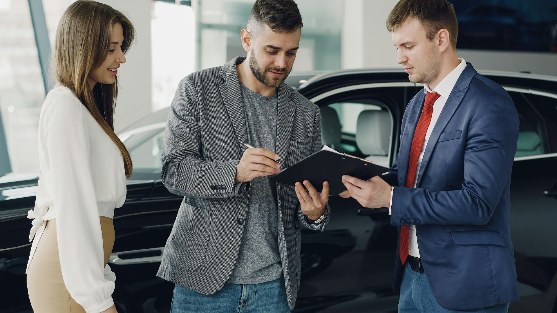 Three adults discussing documents at a car dealership beside a black car.