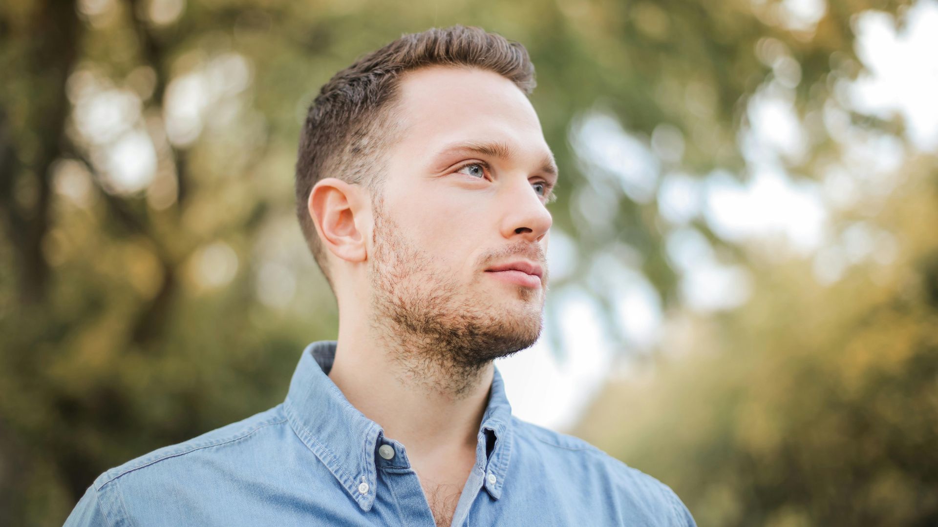 Portrait of a handsome young man with a beard looking thoughtfully in a sunny park setting.