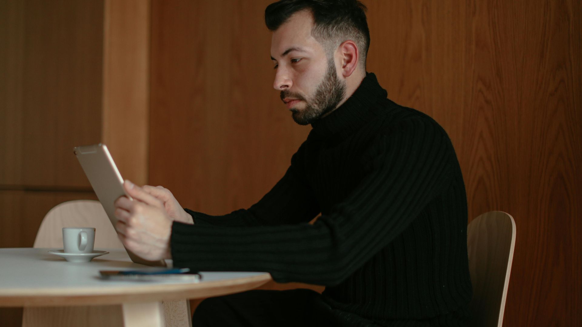 A man in a black sweater uses a tablet in a cozy café, seated at a wooden table.