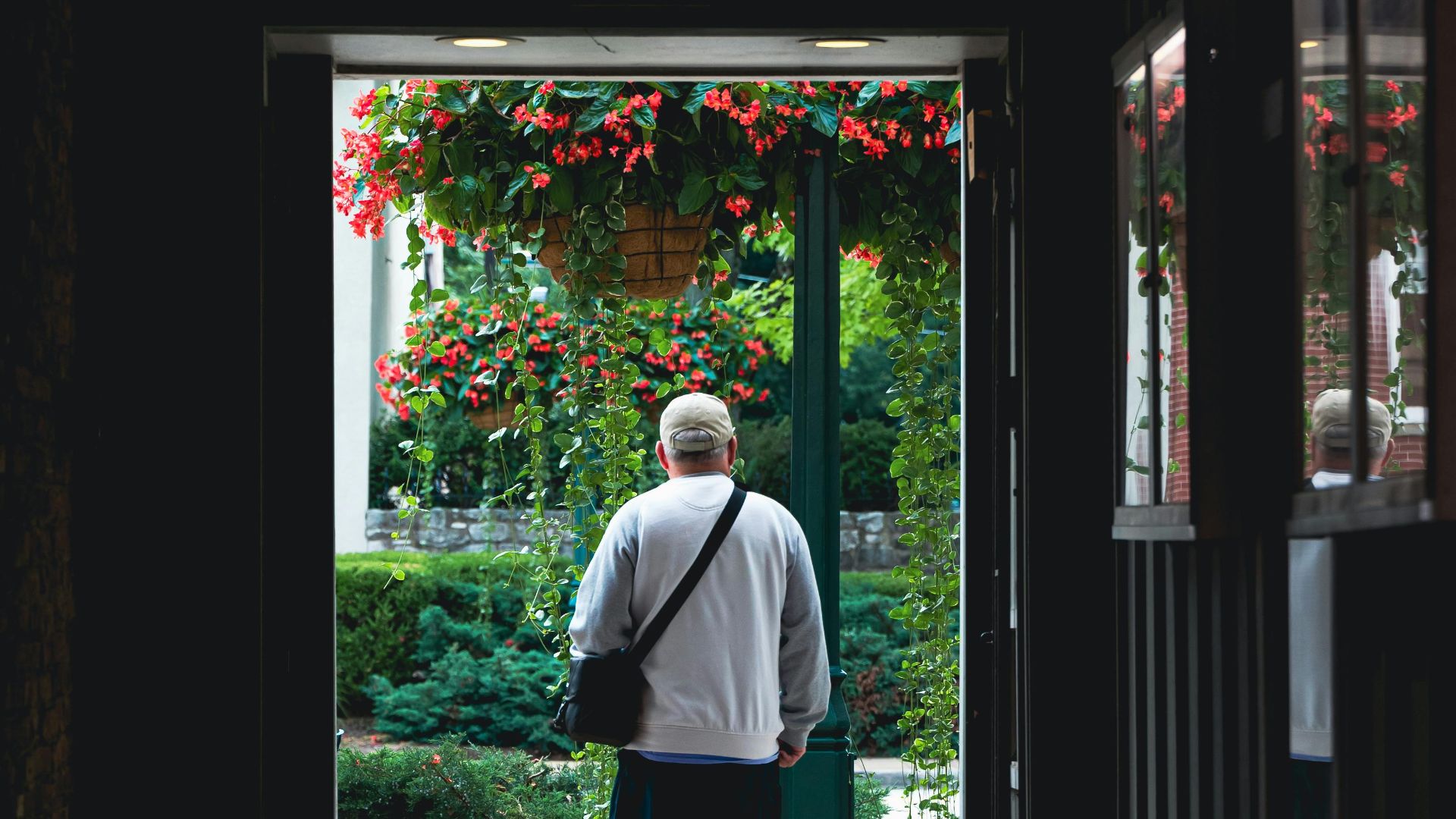 A man walks from a dark hallway into a brightly lit garden area with hanging flowers.