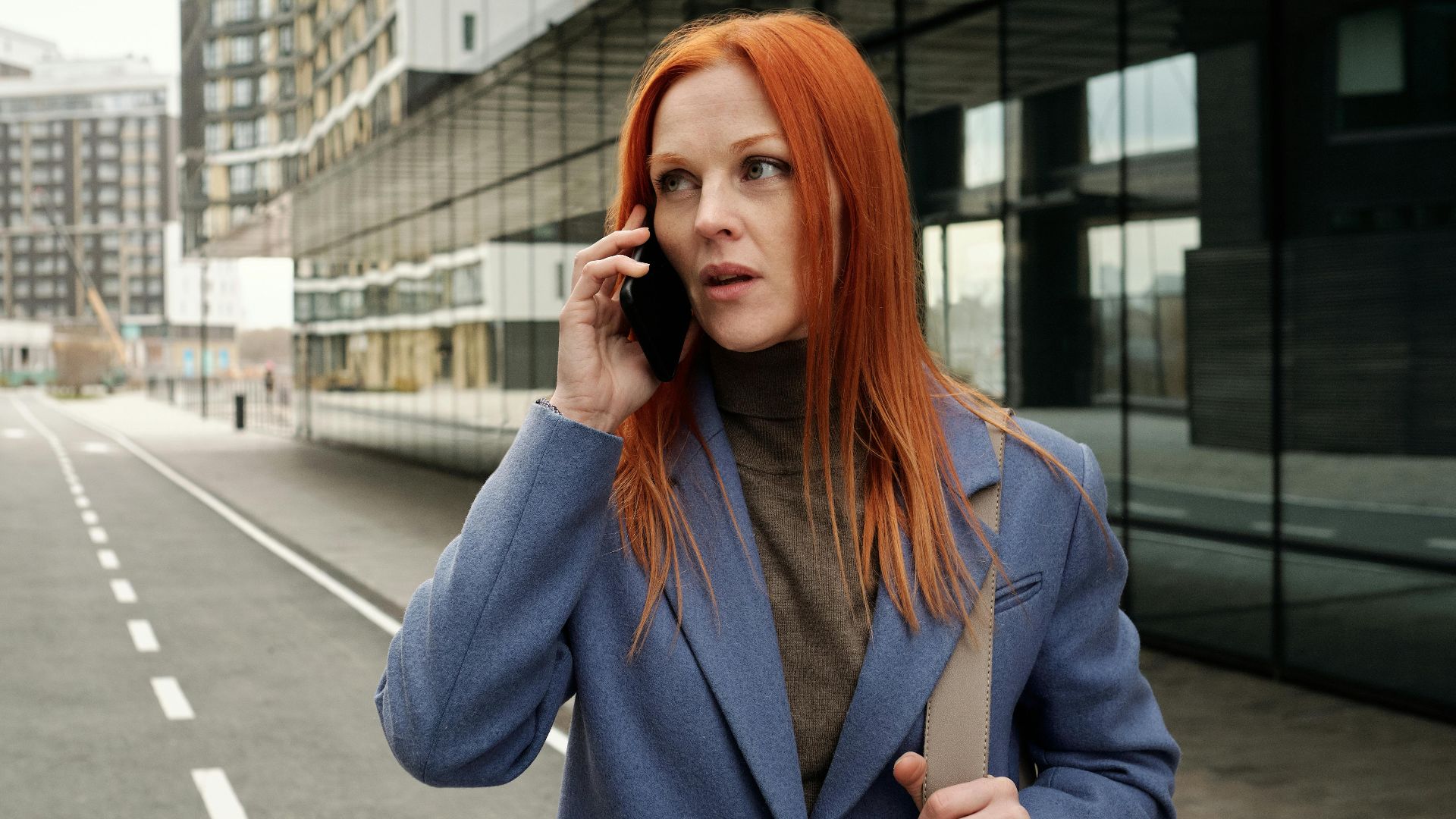 Red-haired woman in blue coat making a phone call on an empty city street.