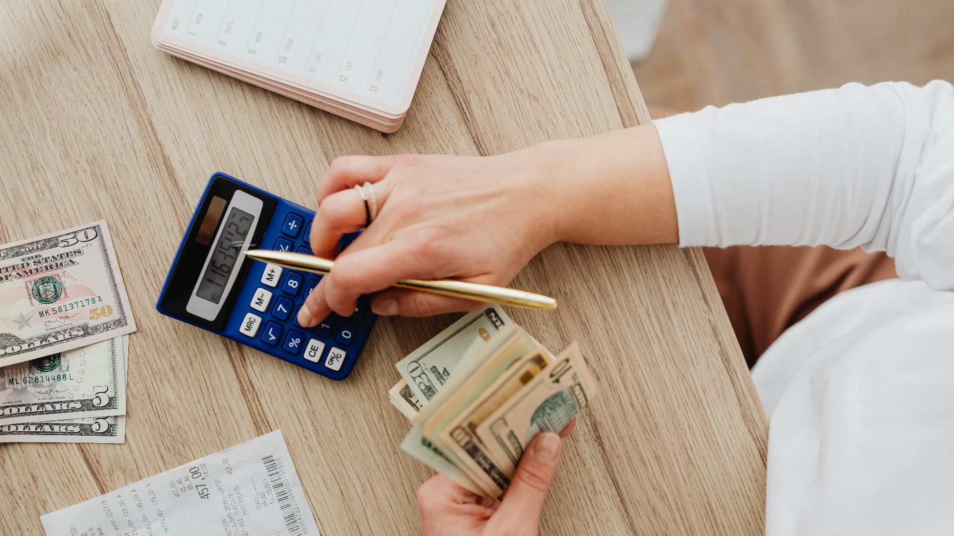 Person using a calculator and counting cash on a wooden table with receipts.