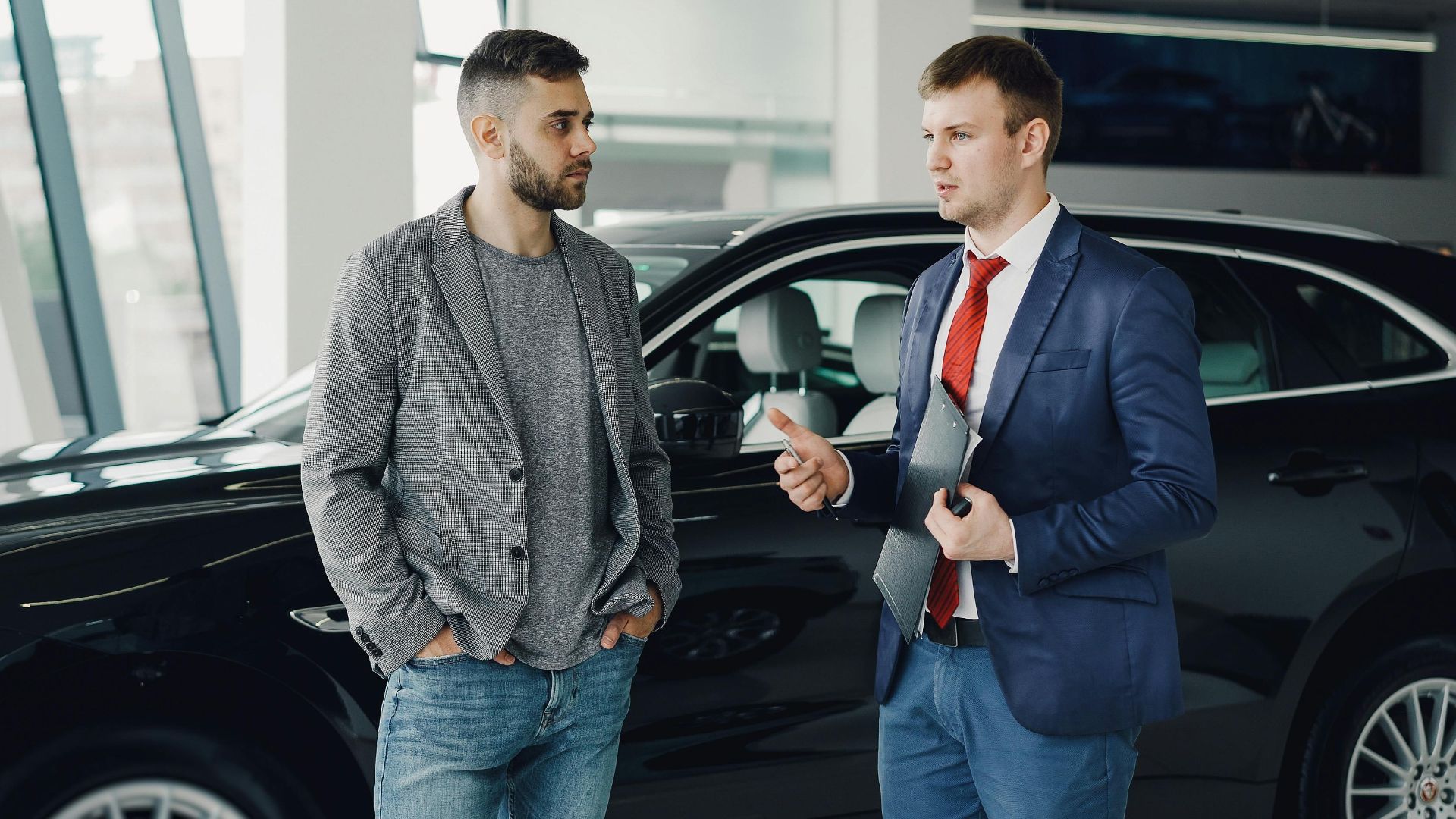 Two men discussing business in a car dealership, standing near a vehicle.