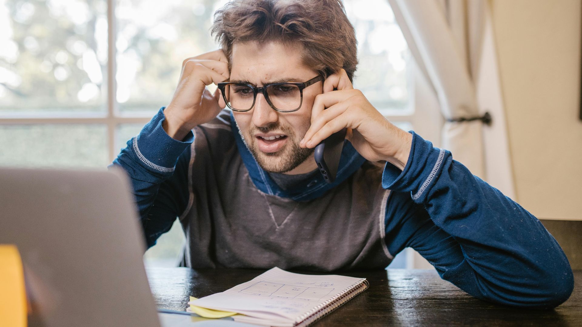 Man in glasses using laptop and smartphone at home, appearing confused.