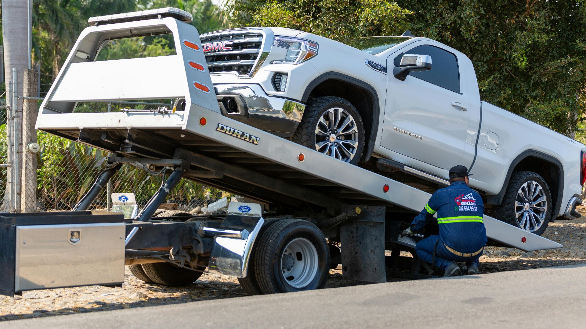 Tow truck operator loading white GMC pickup truck on street in daytime.