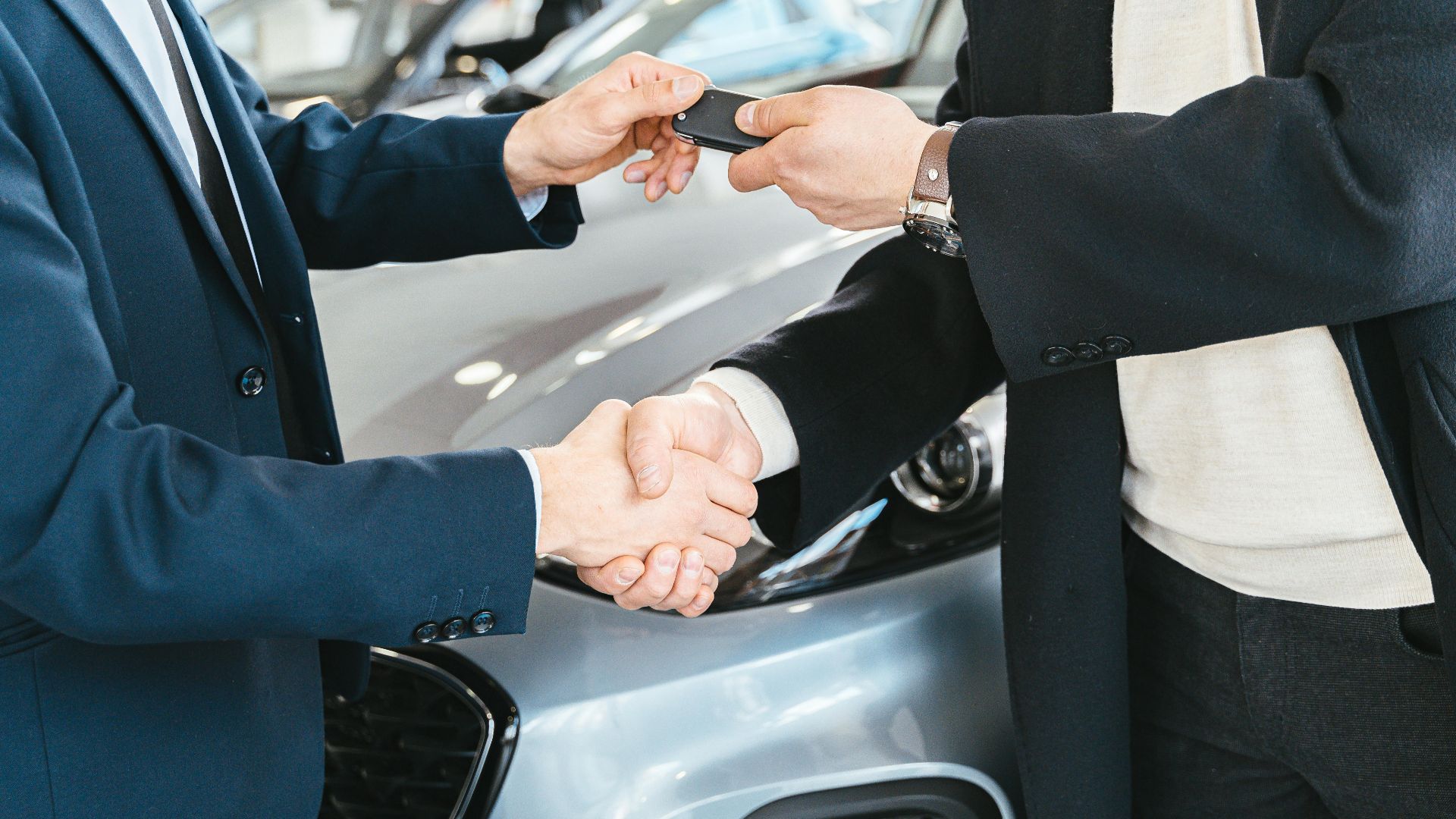 Two businessmen shaking hands and exchanging car keys in a dealership. Symbolizes a successful deal.