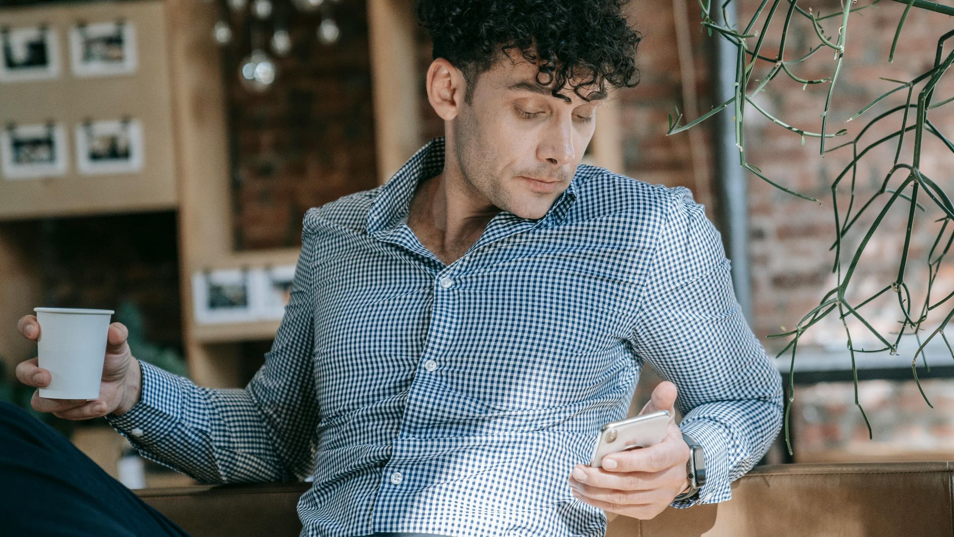 Man in stylish attire enjoying coffee while checking phone in a trendy cafe setting.