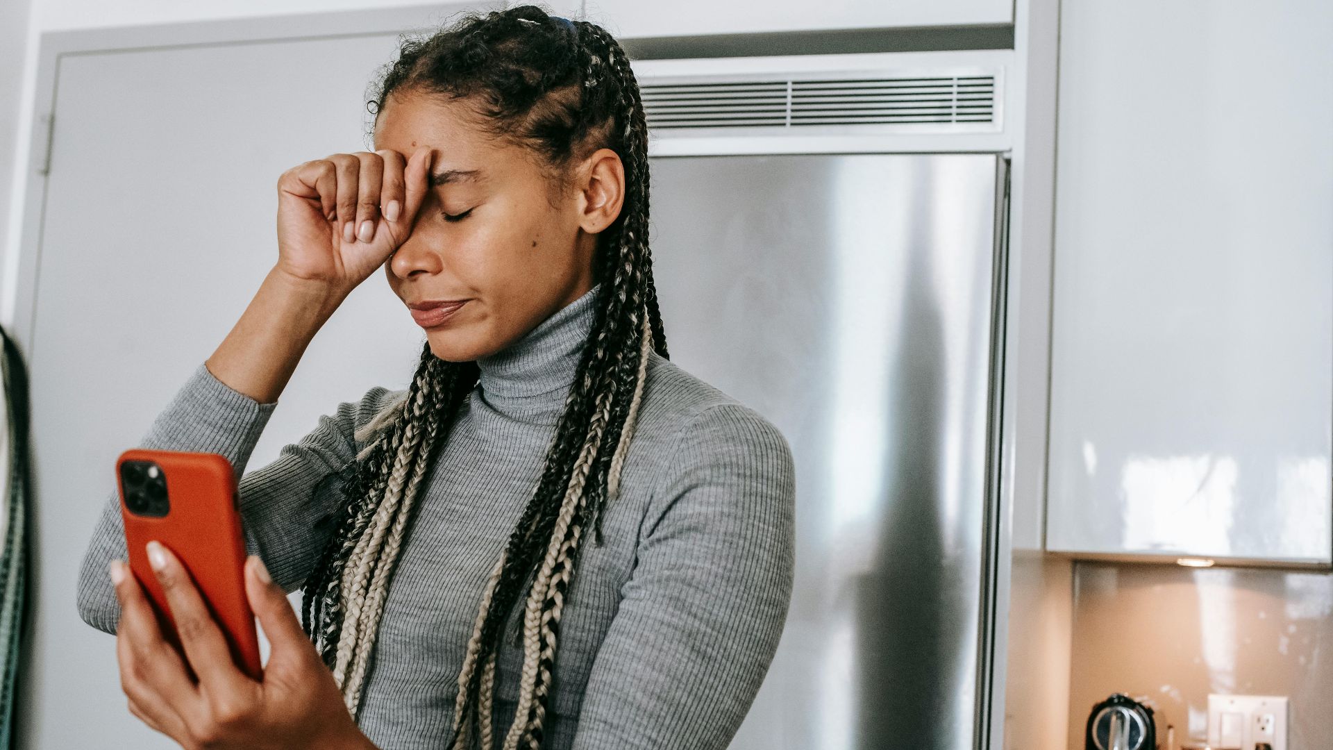Unhappy young African American female with long braids in turtleneck touching face with closed eyes while having video conversation on smartphone in kitchen