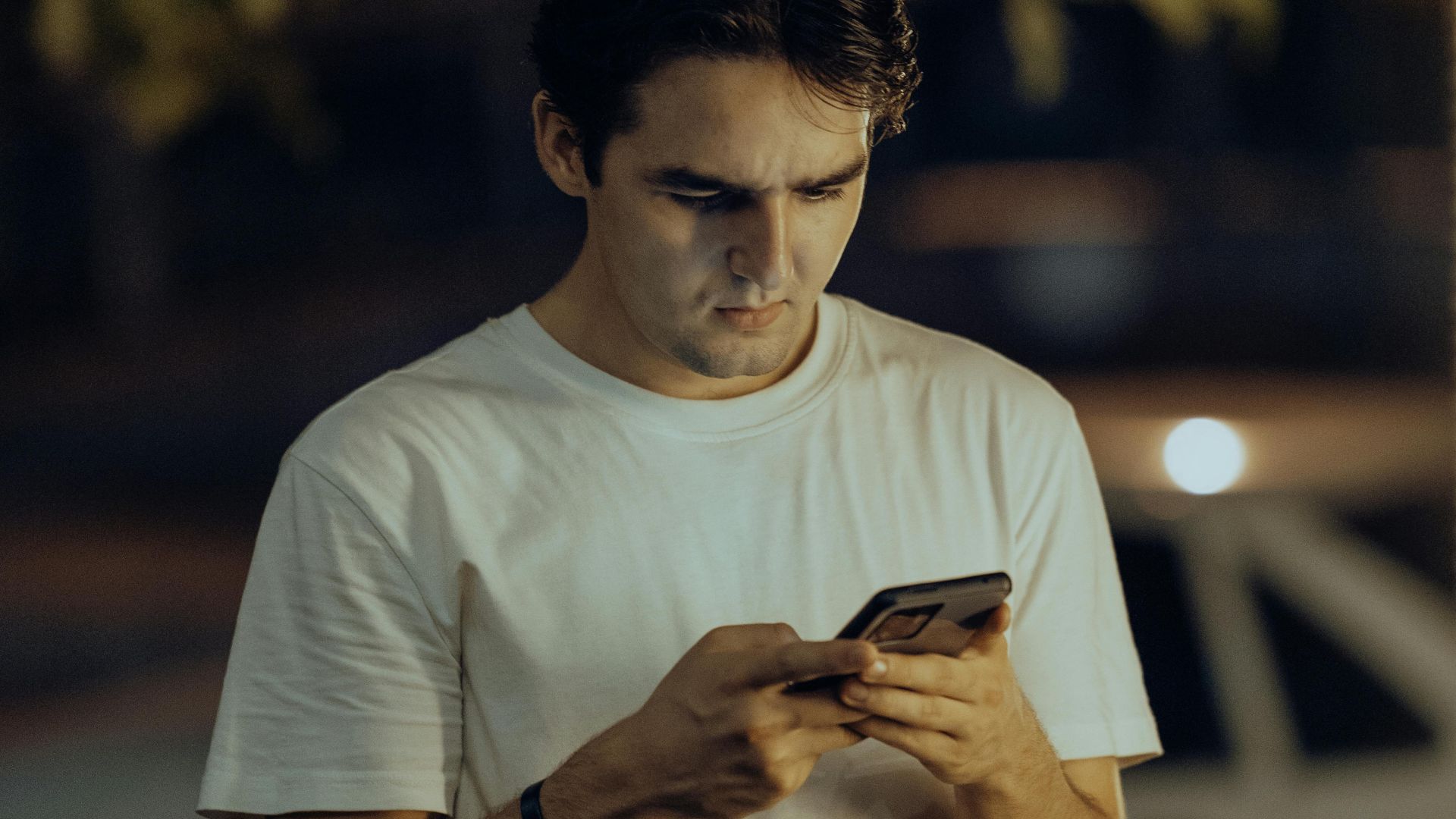 A young man in a white shirt stands outdoors using his smartphone during nighttime.