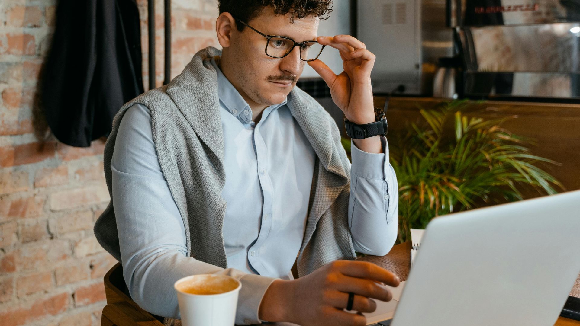 A man with eyeglasses focused on his laptop inside a trendy cafe, enjoying a hot drink.