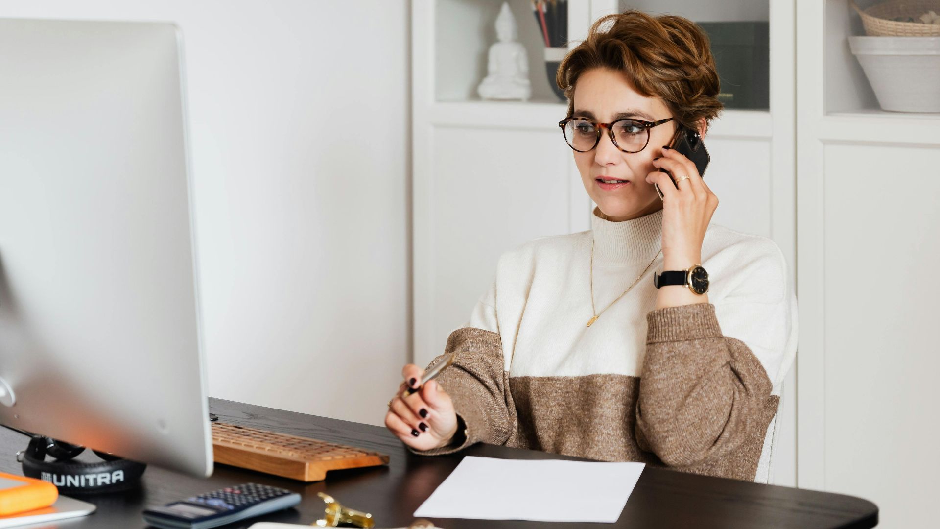 Smart confident female managing director listening to employees report on smartphone while working on computer in light modern office