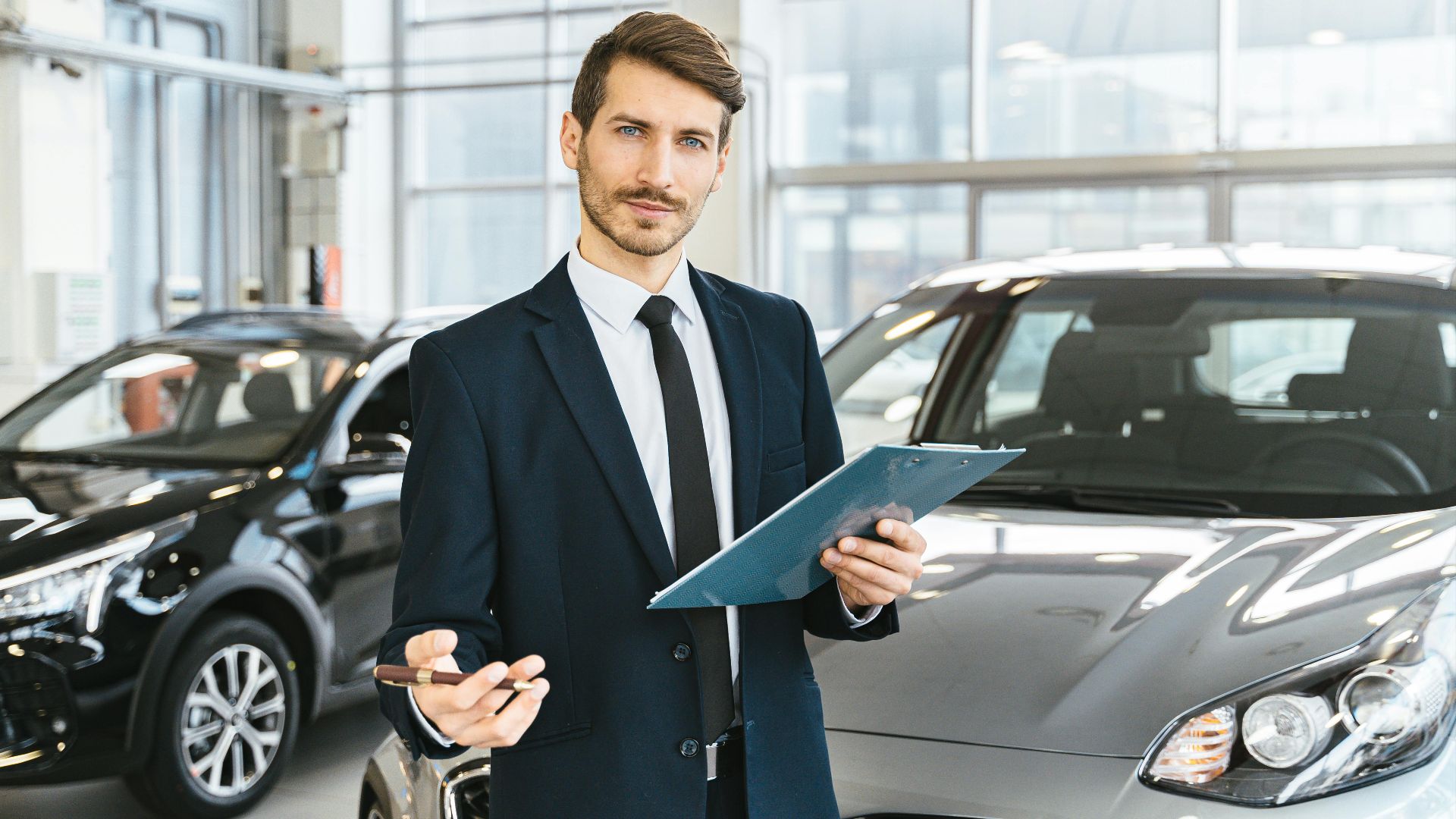 Professional car dealer in business suit holding clipboard in a bright car showroom.
