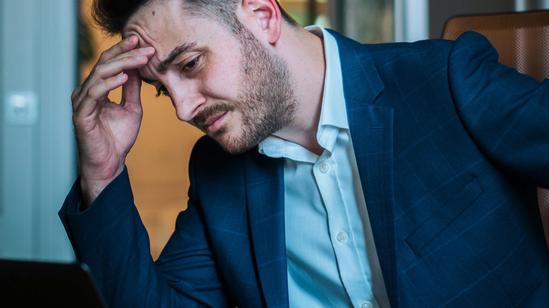 A worried businessman in a blue suit working on his laptop in an office environment.