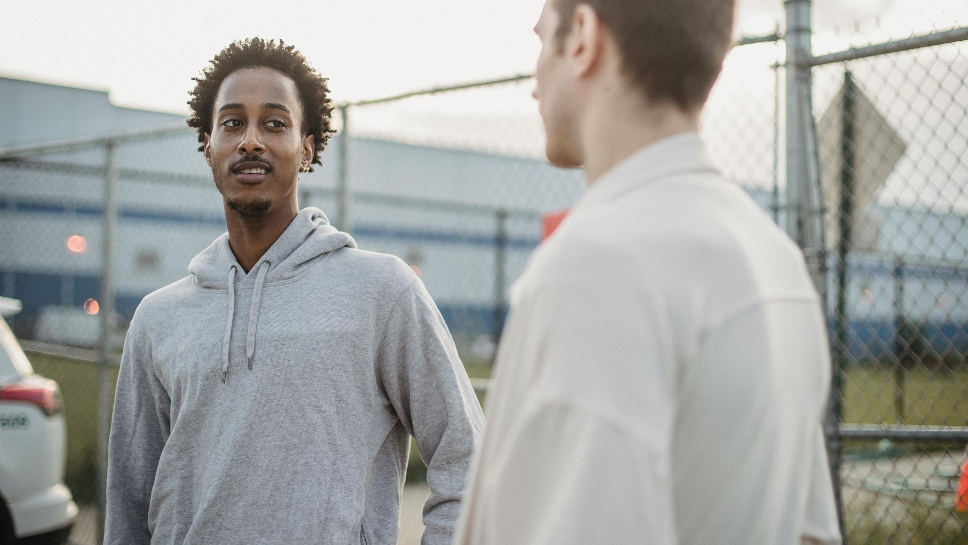 African American male standing with friend against metal grid near car on parking lot