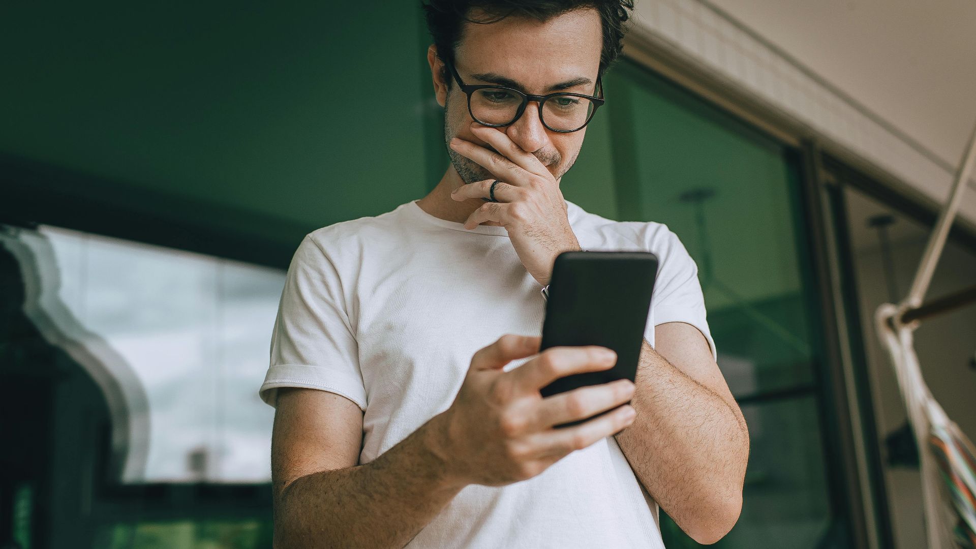A young man in glasses standing on a balcony, using his smartphone with a thoughtful expression.