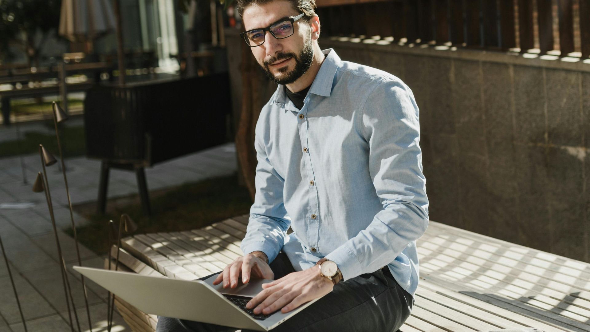A young caucasian man with eyeglasses working on a laptop outdoors on a sunny day.