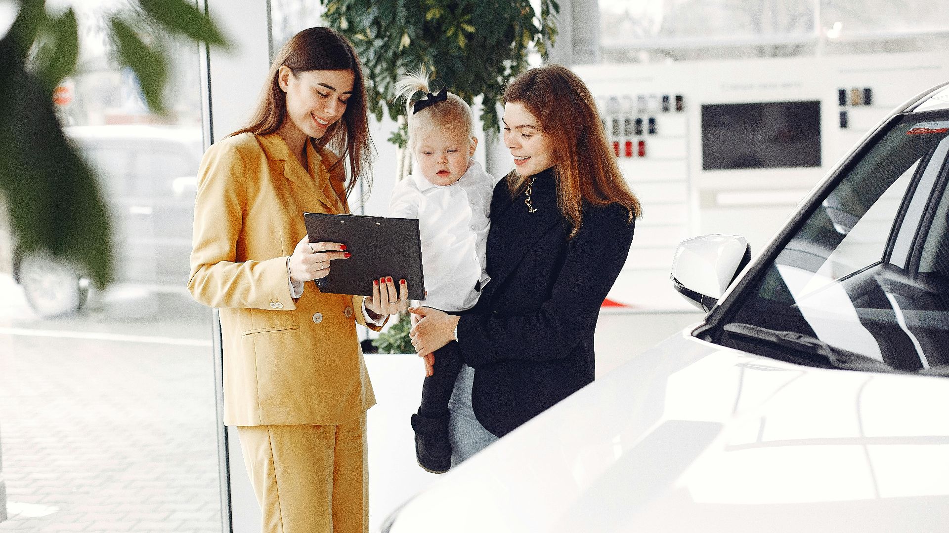Positive young female with cute little daughter on hands discussing car characteristics with professional dealer in stylish beige suit while standing in car showroom in daylight