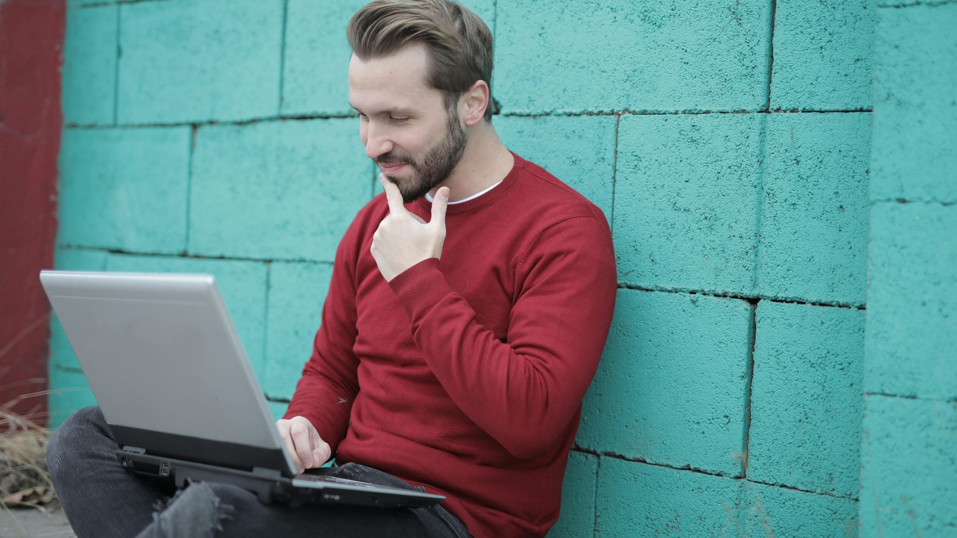 Smiling man using laptop for remote work, leaning on a vibrant green wall outdoors.