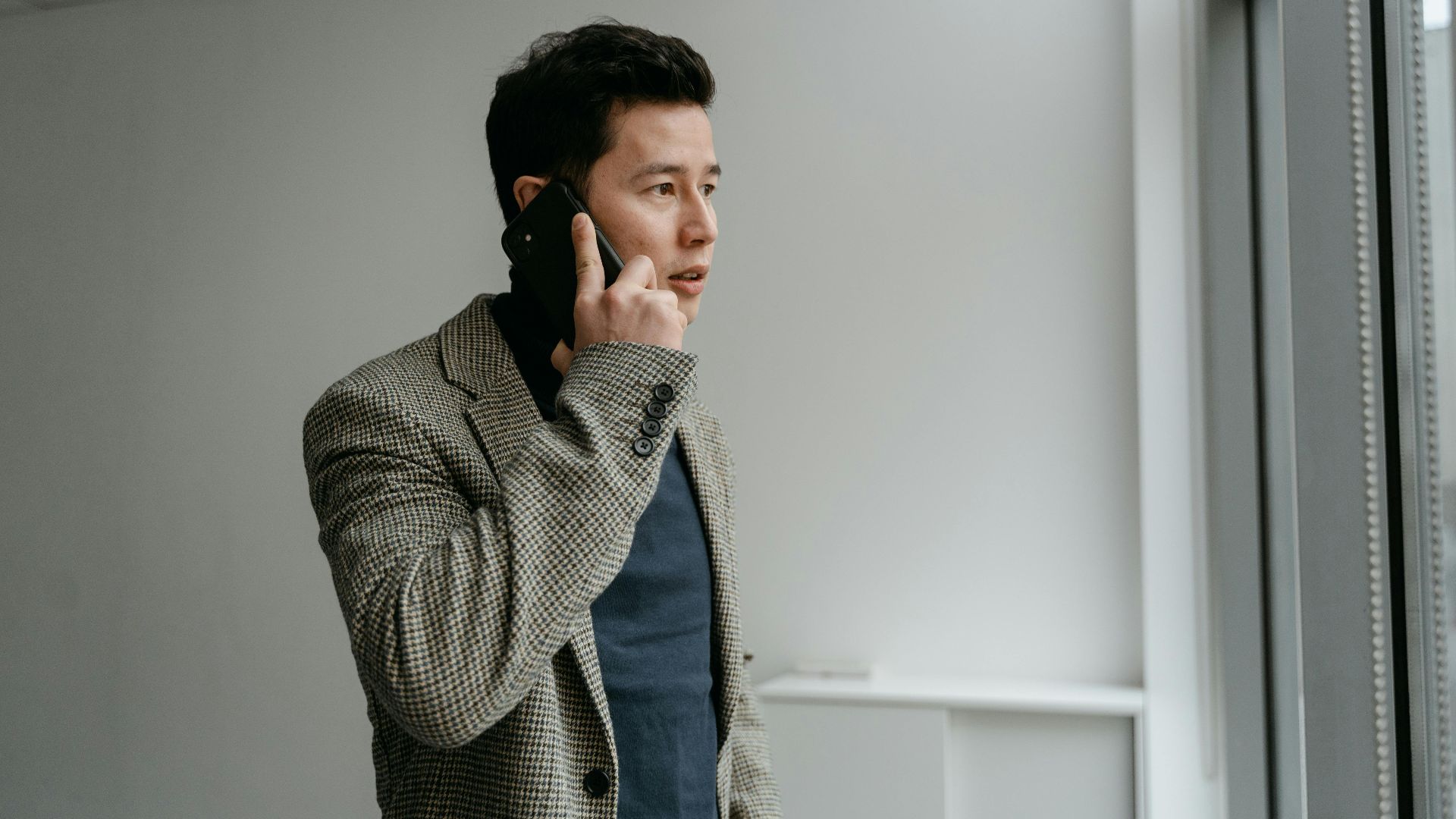 Young businessman in blazer talking on smartphone near window indoors.
