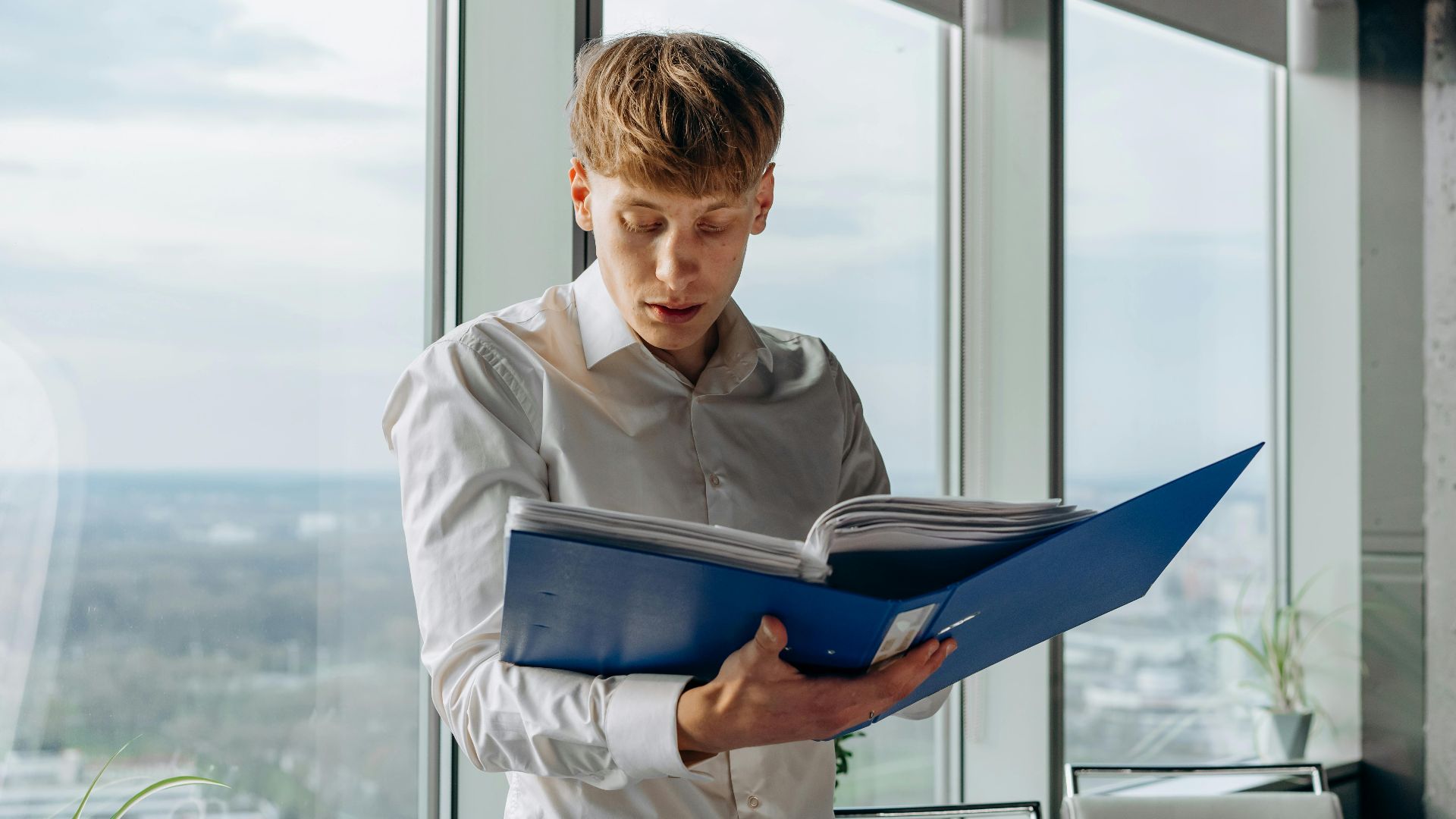 Businessman reading documents in an office with a city view, holding a blue binder.