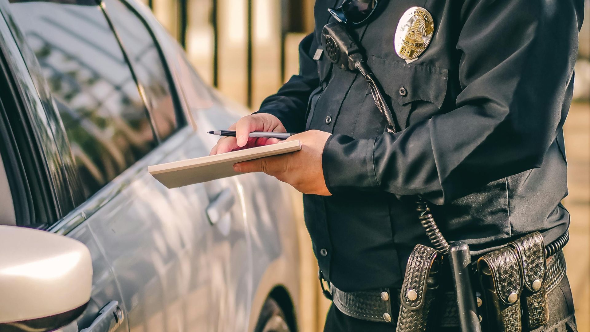 A police officer writes a speeding ticket for a driver in a silver car on a sunny day.