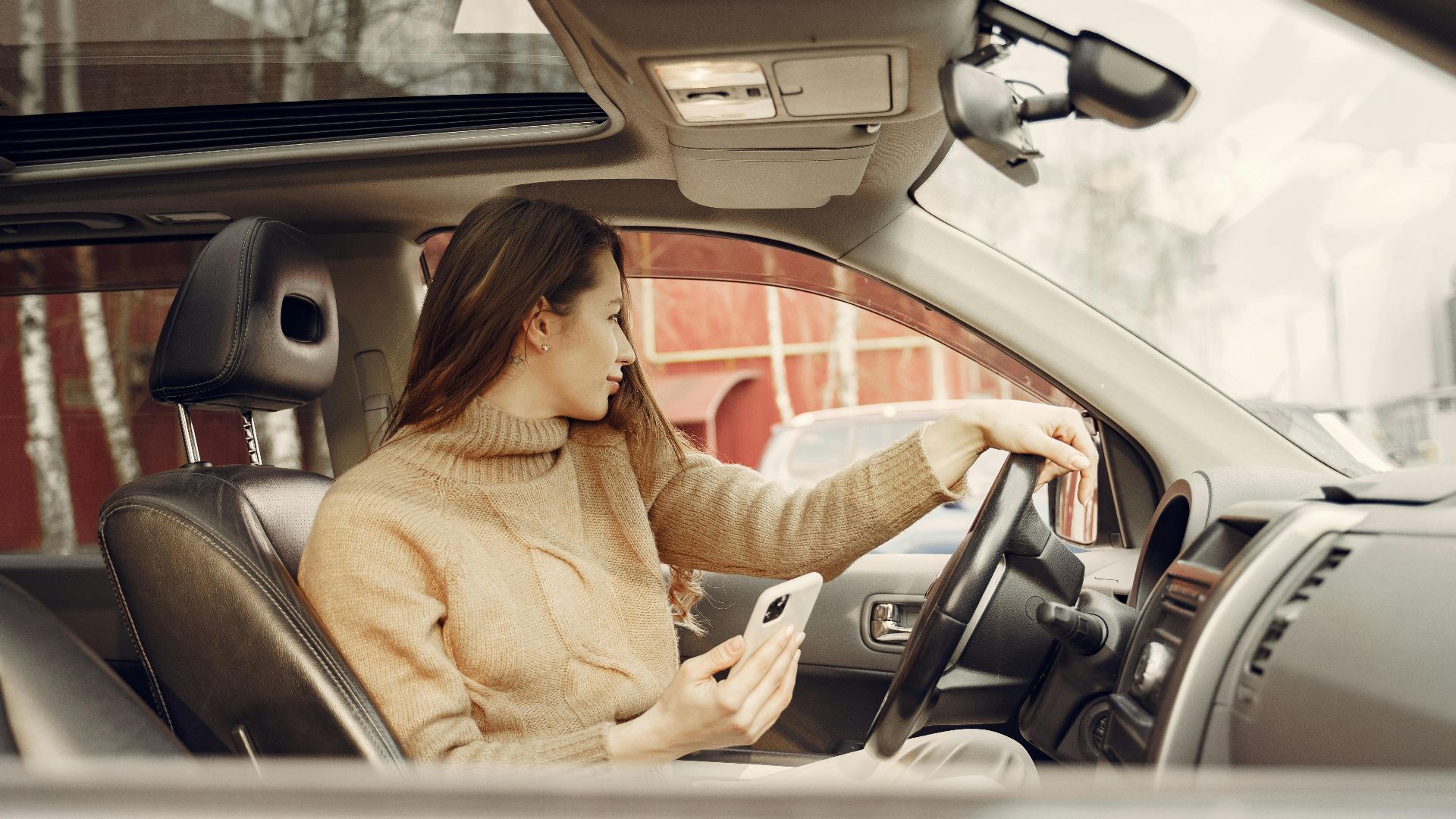 From window side view of adult woman in warm clothes sitting at wheel of modern car waiting for friend and using smartphone