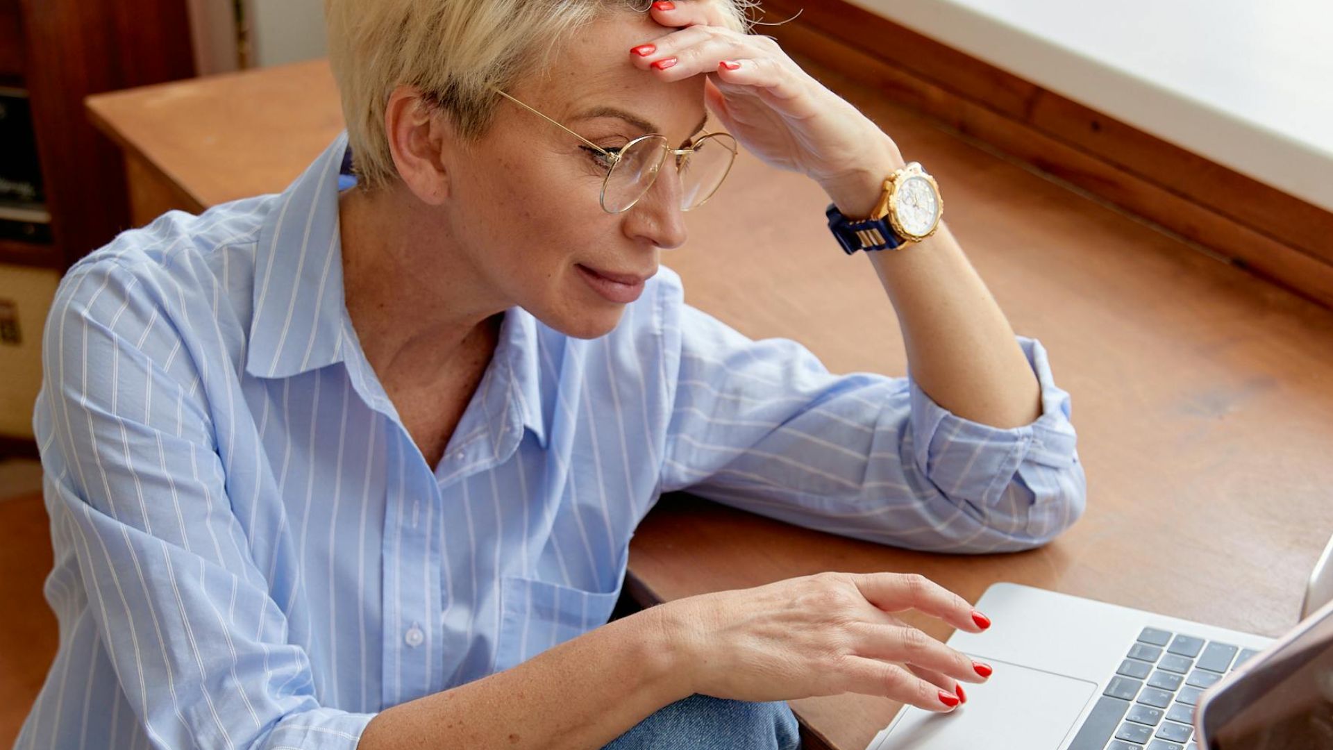 A caucasian woman with glasses using a laptop indoors, sitting by a window.
