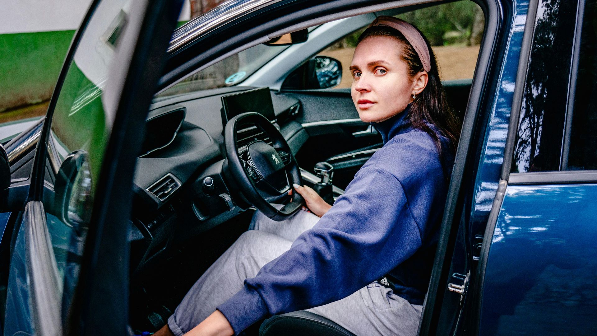 Woman sitting in the driver's seat of a car with an open door, looking outside. Natural setting.