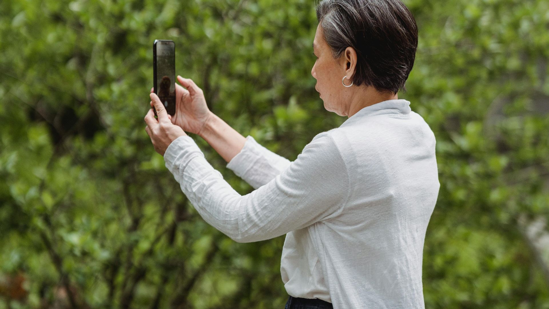 A woman in a white shirt takes a photo with her smartphone in a lush green park.
